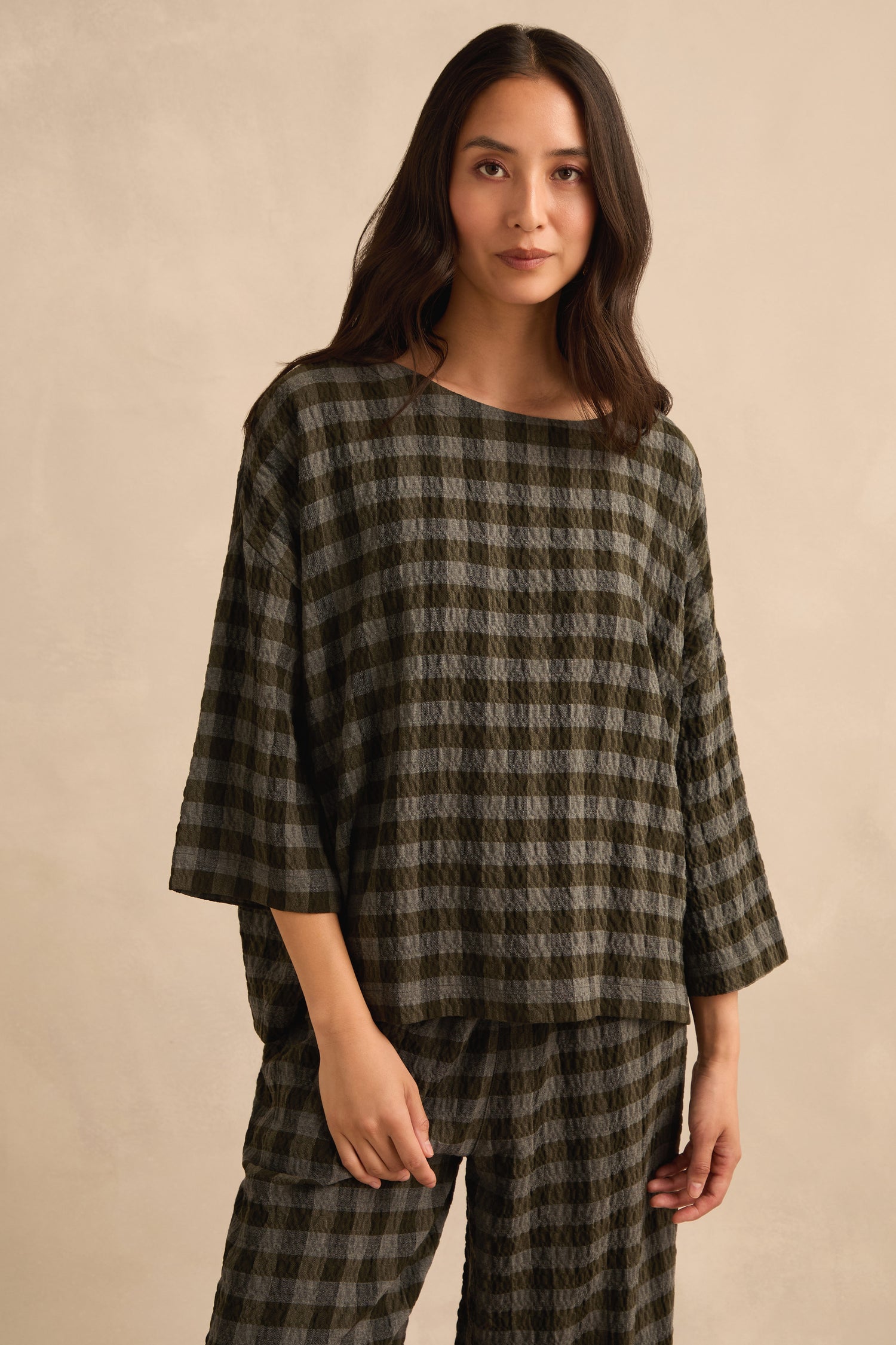 A woman stands against a beige background, facing forward with one hand by her side, wearing the Crinkle Check Boxy Top and matching pants in an oversized, loose fit.