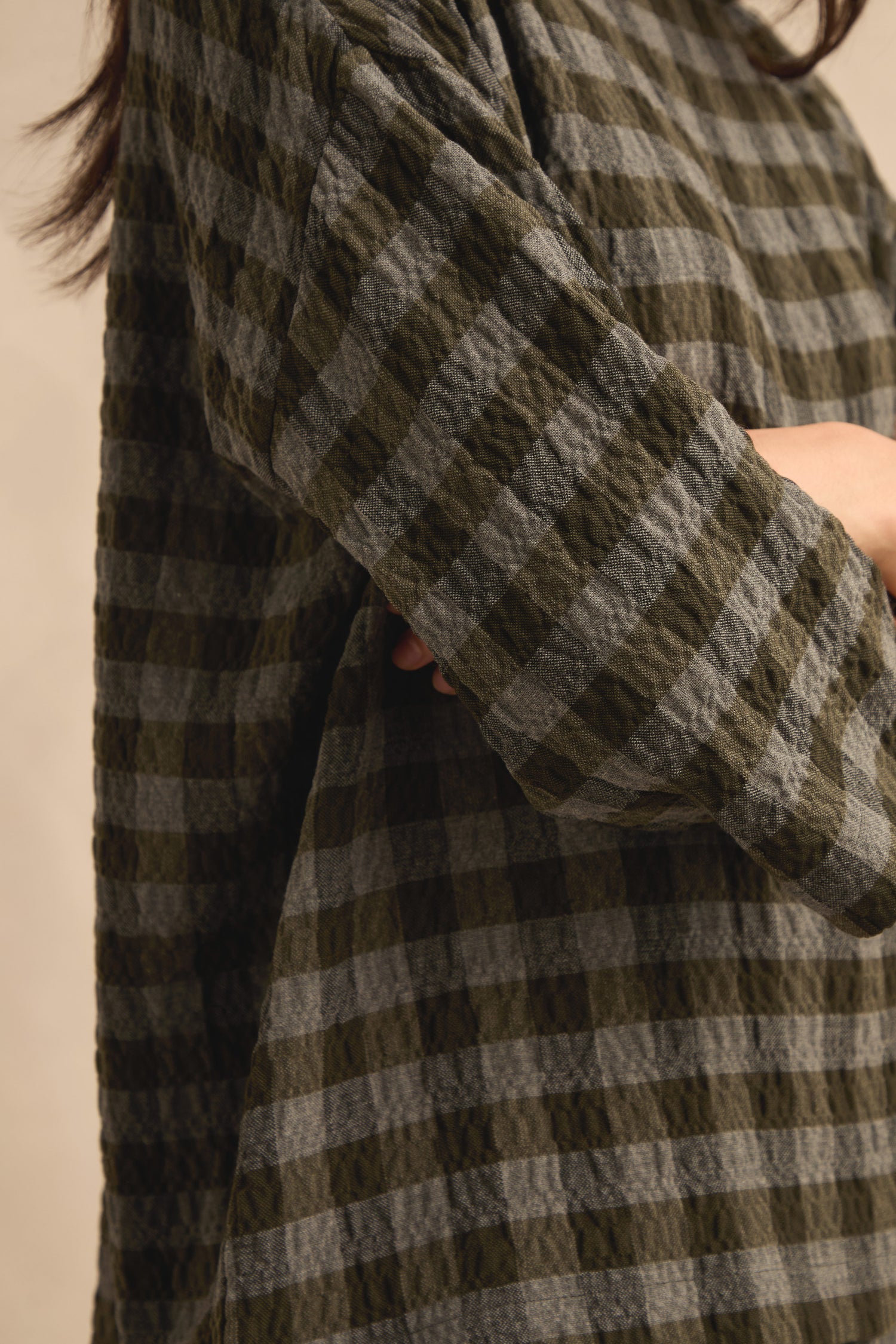 Close-up of a person wearing the Crinkle Check Boxy Top, a textured long-sleeve shirt in green and gray plaid with an oversized fit. The individual’s arms are folded, and only part of the upper body is visible.