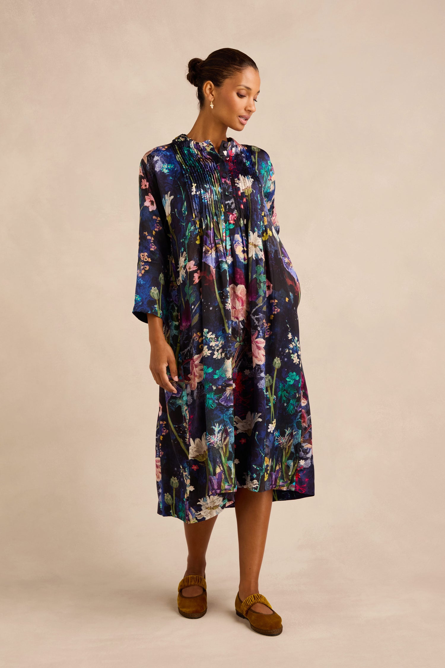 Wearing the Floral Bouquet Pleated Dress, a woman poses against a neutral background. The dark, pleated midi-length dress features long sleeves and an all-over floral pattern, styled with brown moccasins and her hair in a bun. Crafted through artisan production, this elegant cupro dress showcases delicate pleats and thoughtful details.