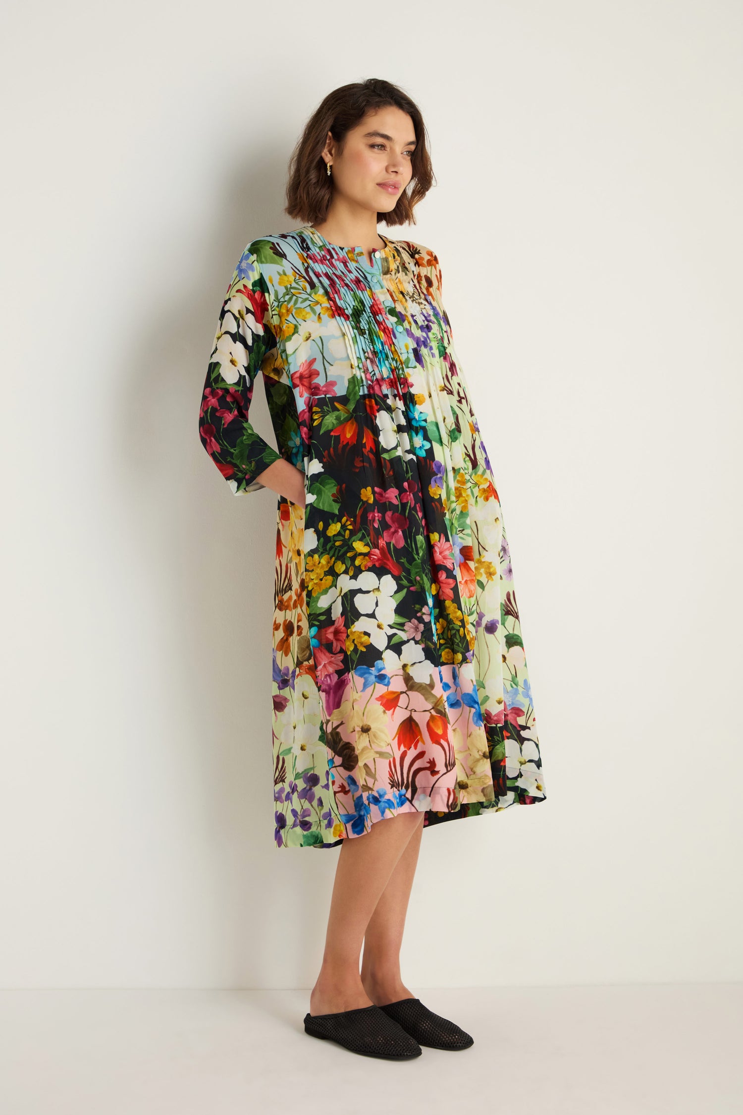 Woman stands against a plain wall wearing the Impressionist Garden Pleated Dress, a colorful, long-sleeved, floral print midi dress with pleats, paired with black slip-on shoes.