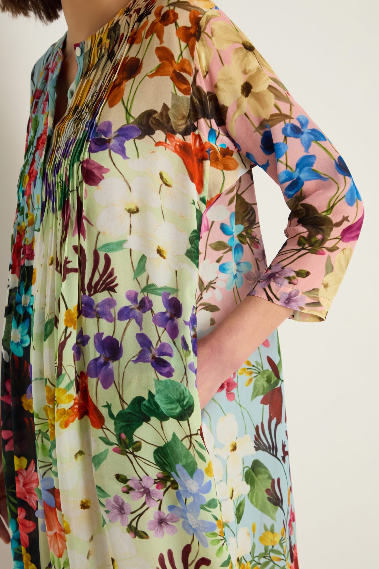 Wearing the Impressionist Garden Pleated Dress, a person stands with one hand in their pocket against a plain background.