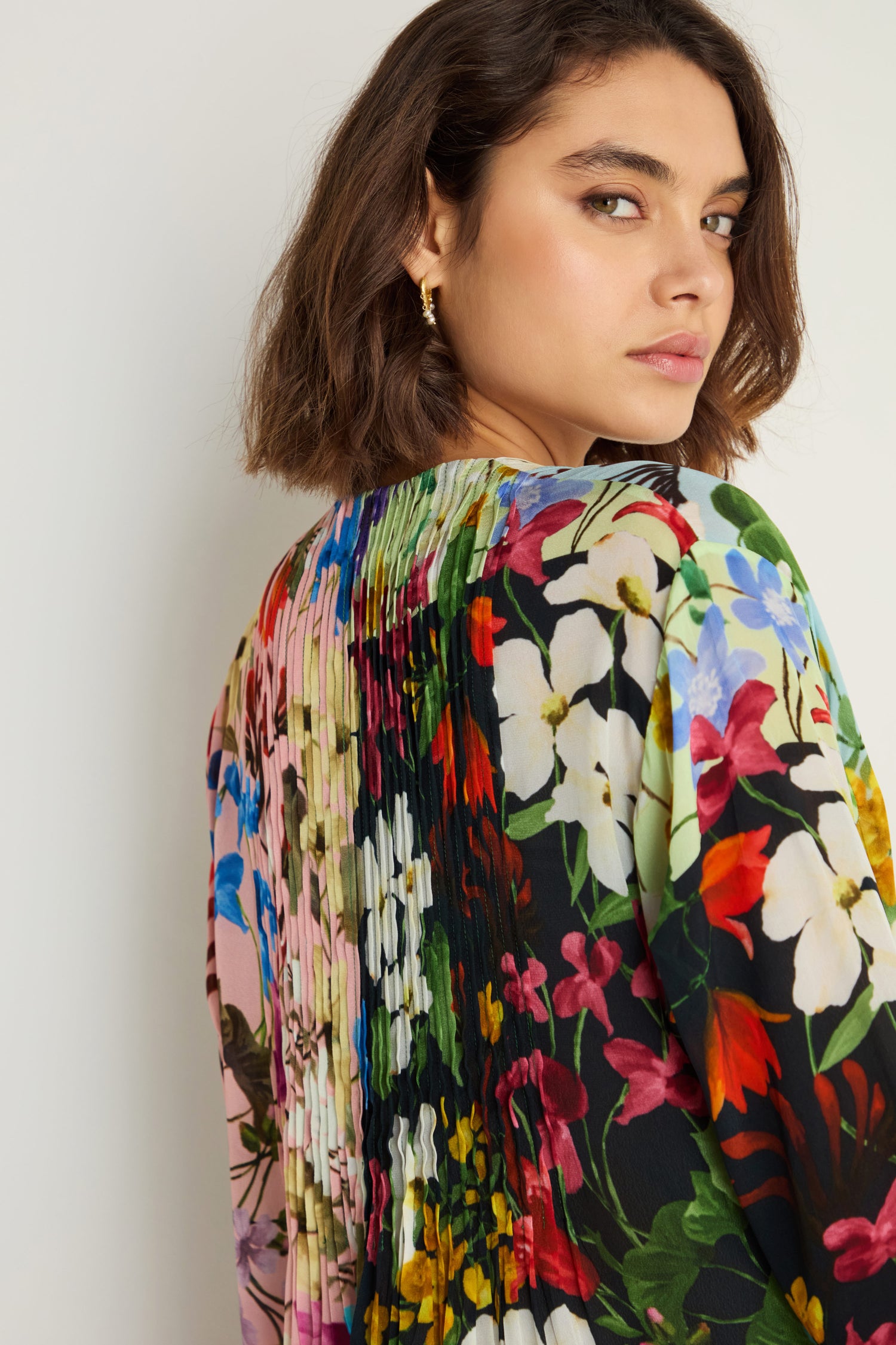 A woman with short brown hair looks over her shoulder, wearing the Impressionist Garden Pleated Dress—a multi-colored floral print—against a light background.