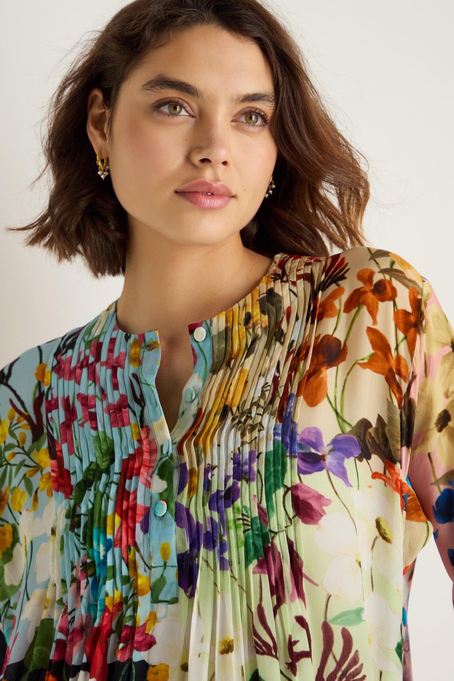 Woman with short brown hair wears the Impressionist Garden Pleated Dress, featuring a colorful floral print and pleats, standing against a plain light background.