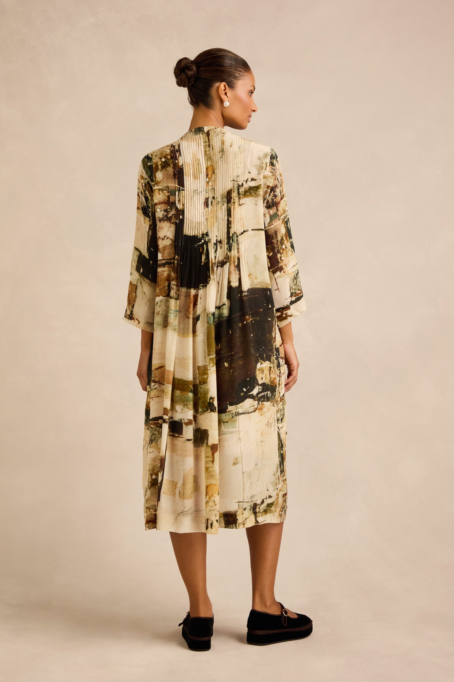 Woman facing away, wearing a loose, long-sleeved Block Print Pleated Dress with an abstract beige, black, and brown pattern and black slip-on shoes, standing against a plain light background.