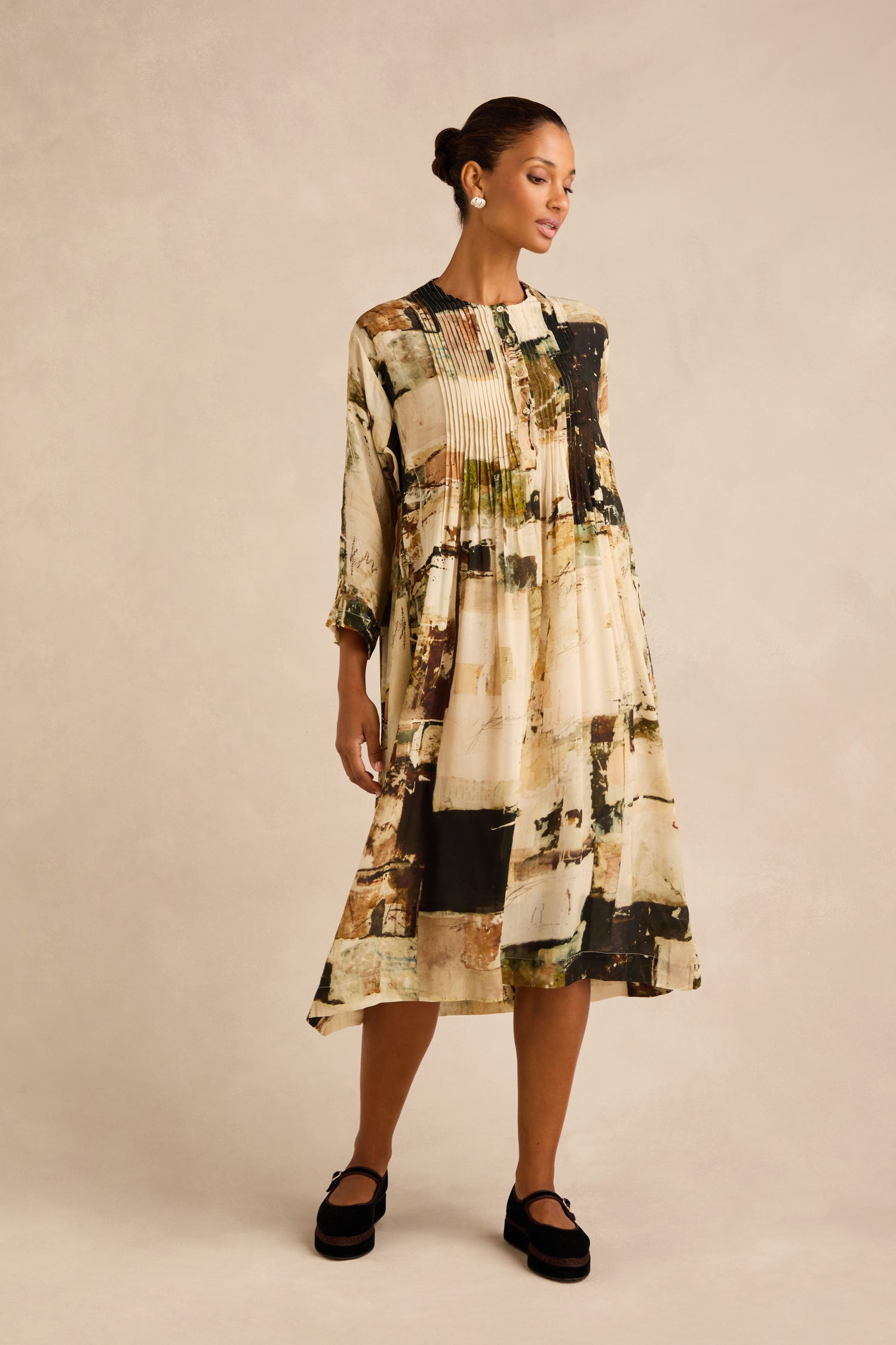 A woman wearing the Block Print Pleated Dress—a long-sleeved, pleated midi dress crafted from luxurious cupro viscose—styled with black shoes and earrings, stands against a neutral background.