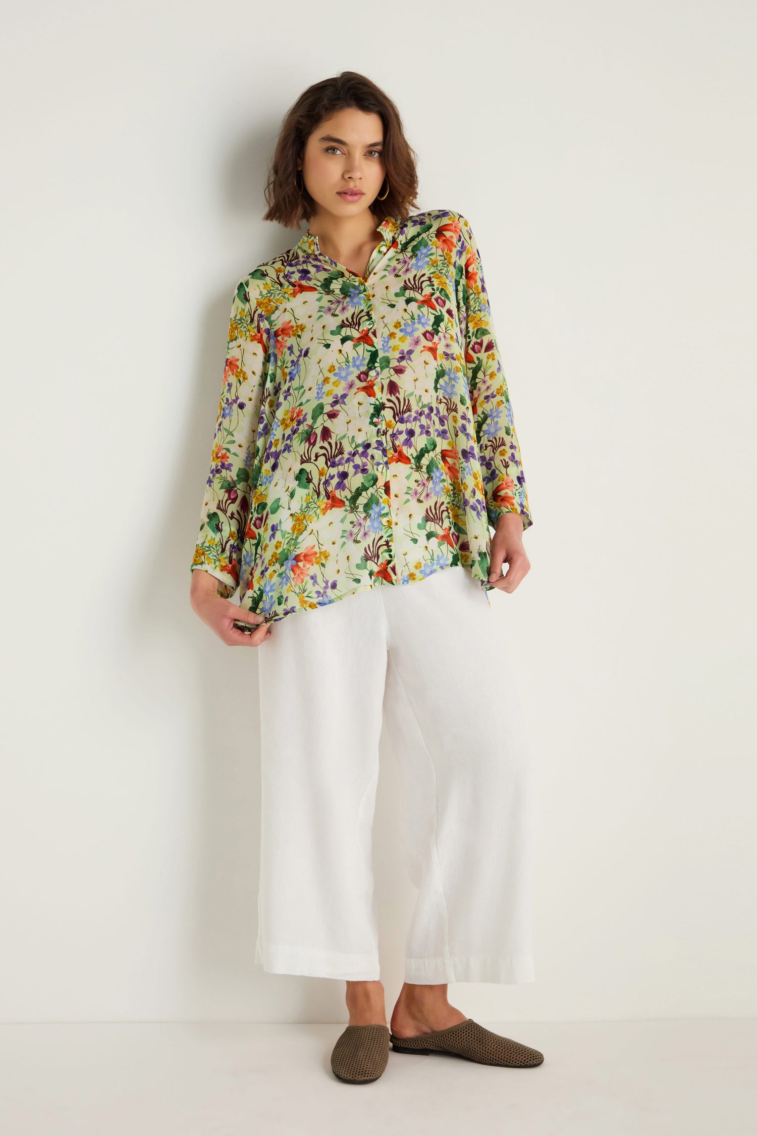 A woman stands against a white background wearing the Country Garden Cupro Shirt with long sleeves, floral pattern, white wide-leg pants, and brown slip-on shoes.