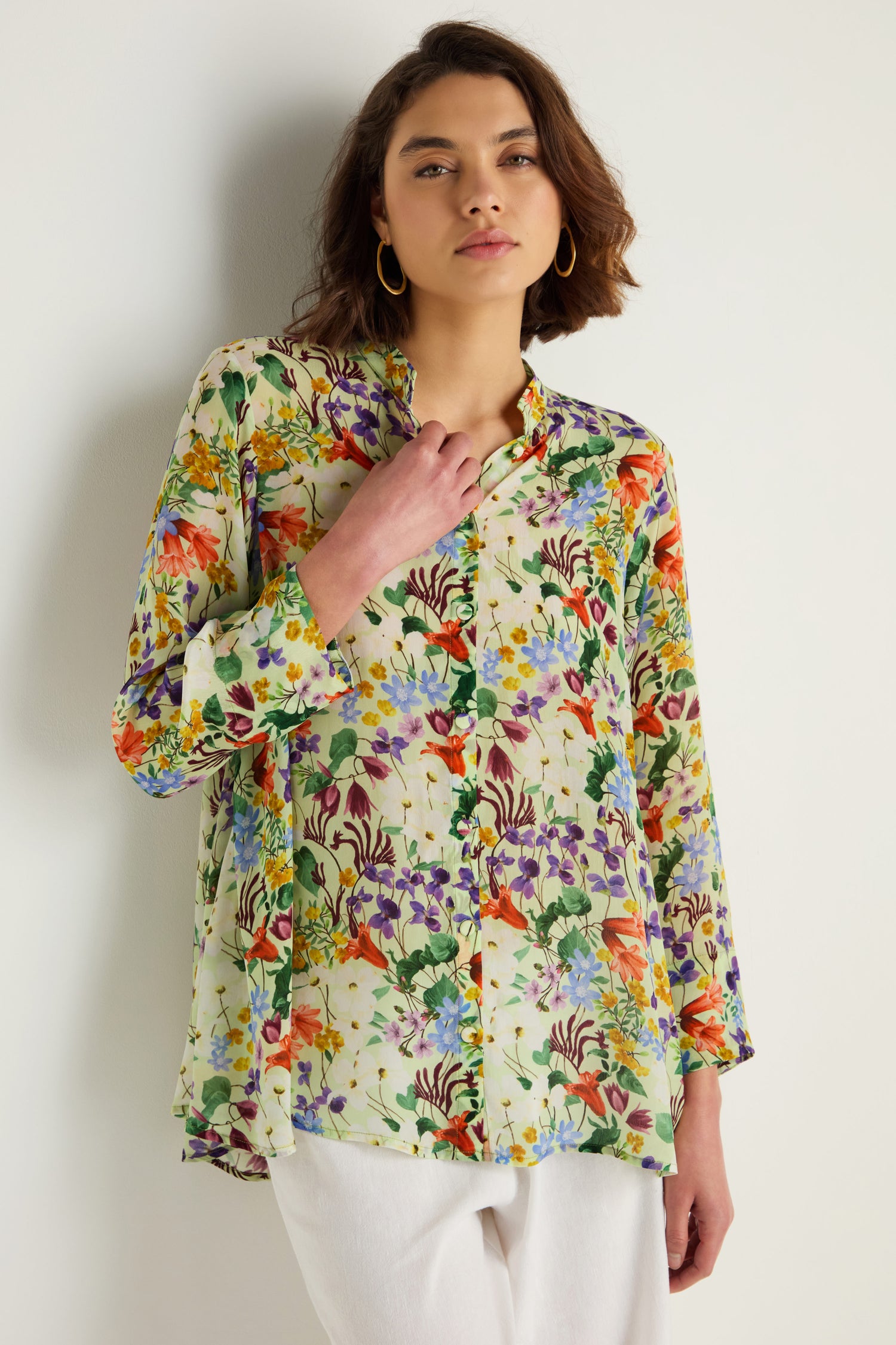 Wearing the sheer, long-sleeve Country Garden Cupro Shirt with a colorful floral pattern and white pants, the person stands against a plain light background.