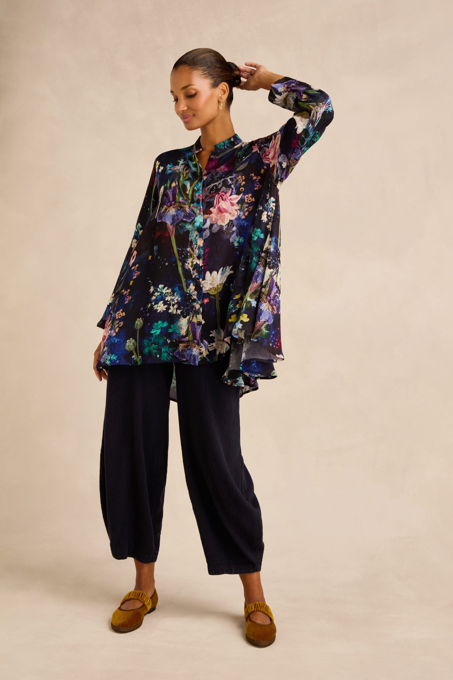 A woman stands against a neutral background wearing the Floral Bouquet Flared Shirt, crafted from a cupro viscose blend in a loose, swing silhouette. She pairs it with wide-leg black pants and yellow shoes, her hair pulled back and one arm raised.