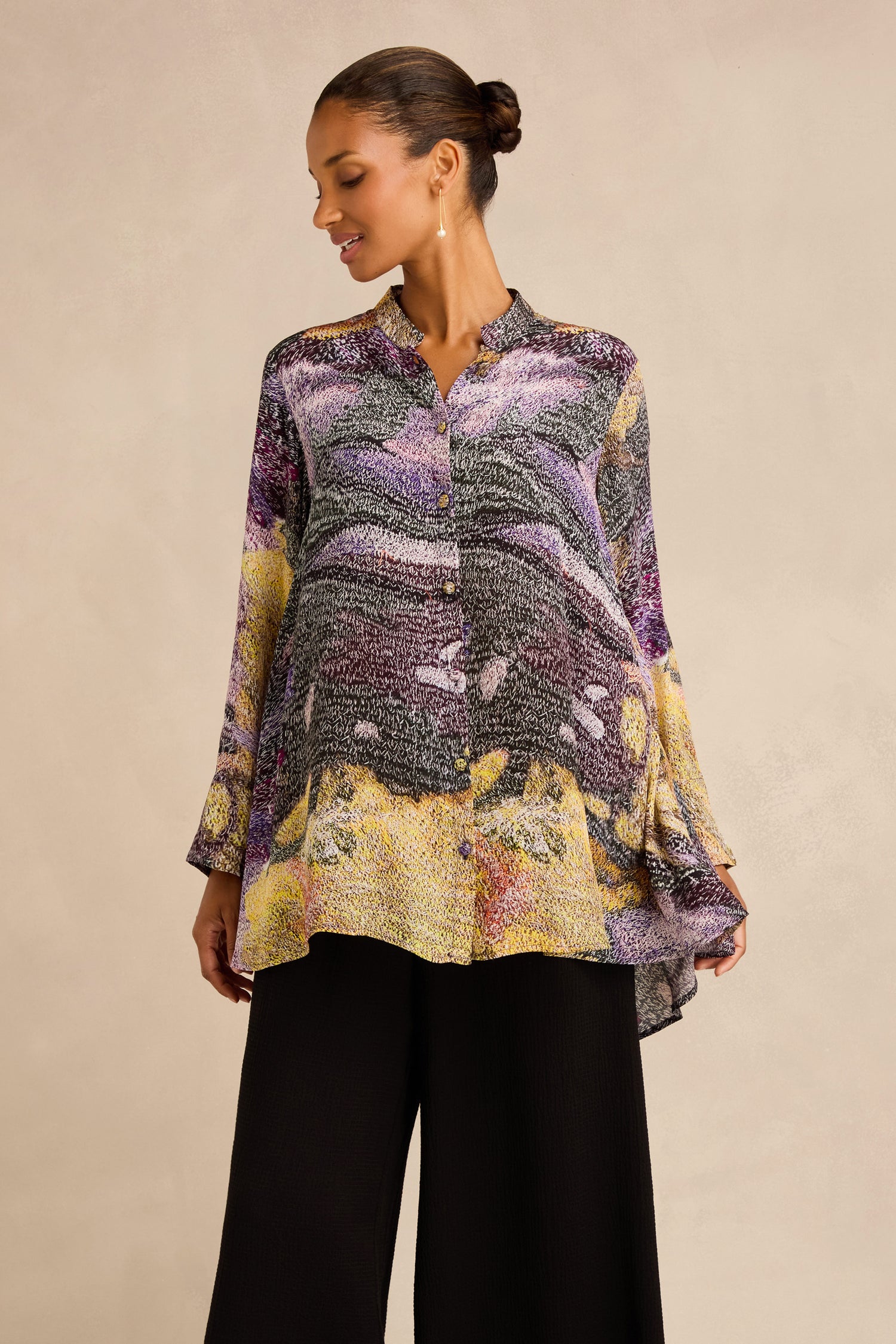 A woman models the Impressionist Print Flared Shirt, a loose, long-sleeved blouse crafted from a luxurious blend of cupro and viscose. Showcasing abstract patterns in purple, yellow, and black, this artisan-produced top is paired with wide-leg black pants. She stands against a plain beige background.