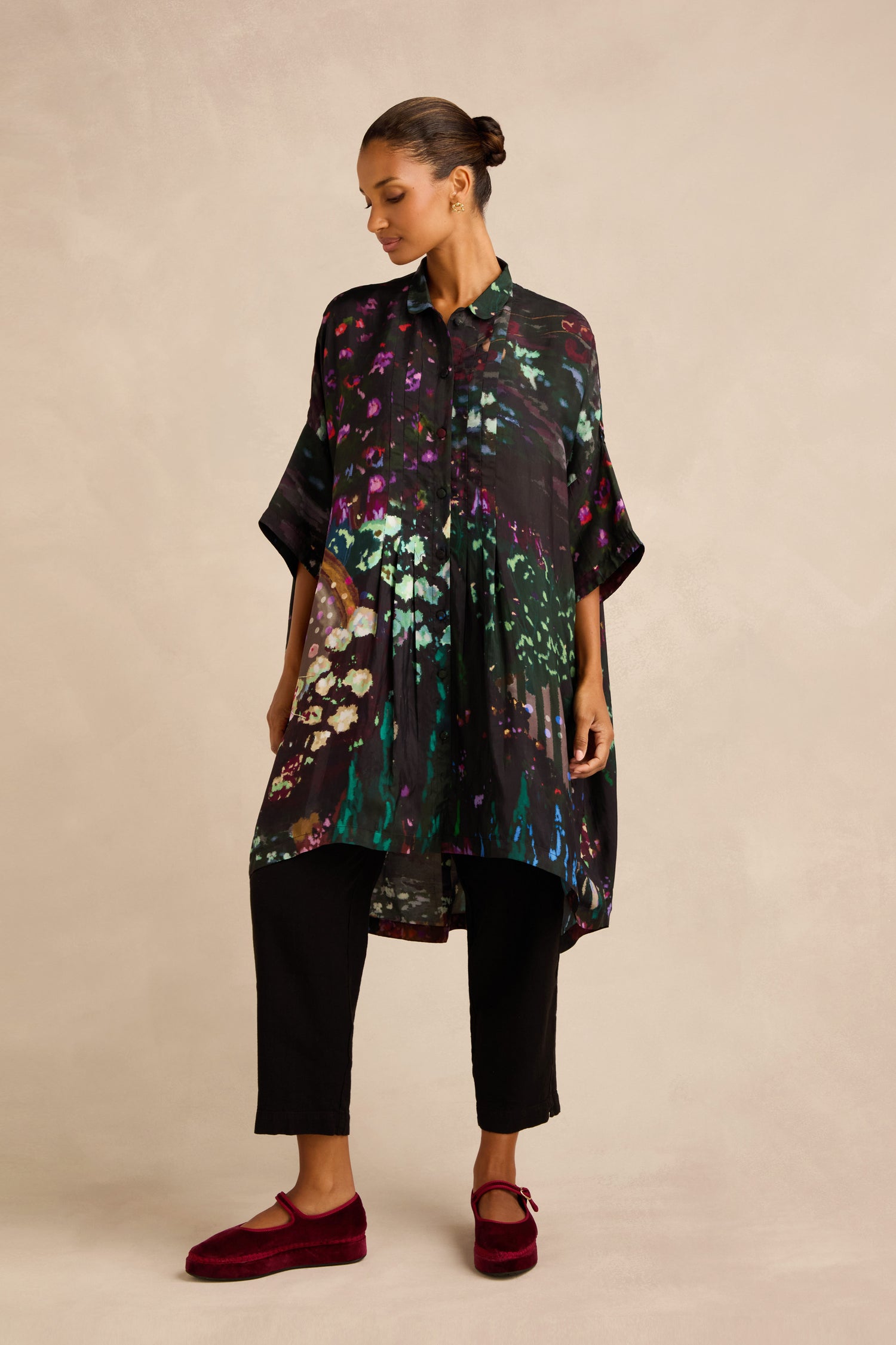 A woman wears the Water Lilies Button Through Tunic with a loose floral print over black pants and burgundy flats, standing against a neutral background—a look that beautifully showcases artisan craftsmanship.