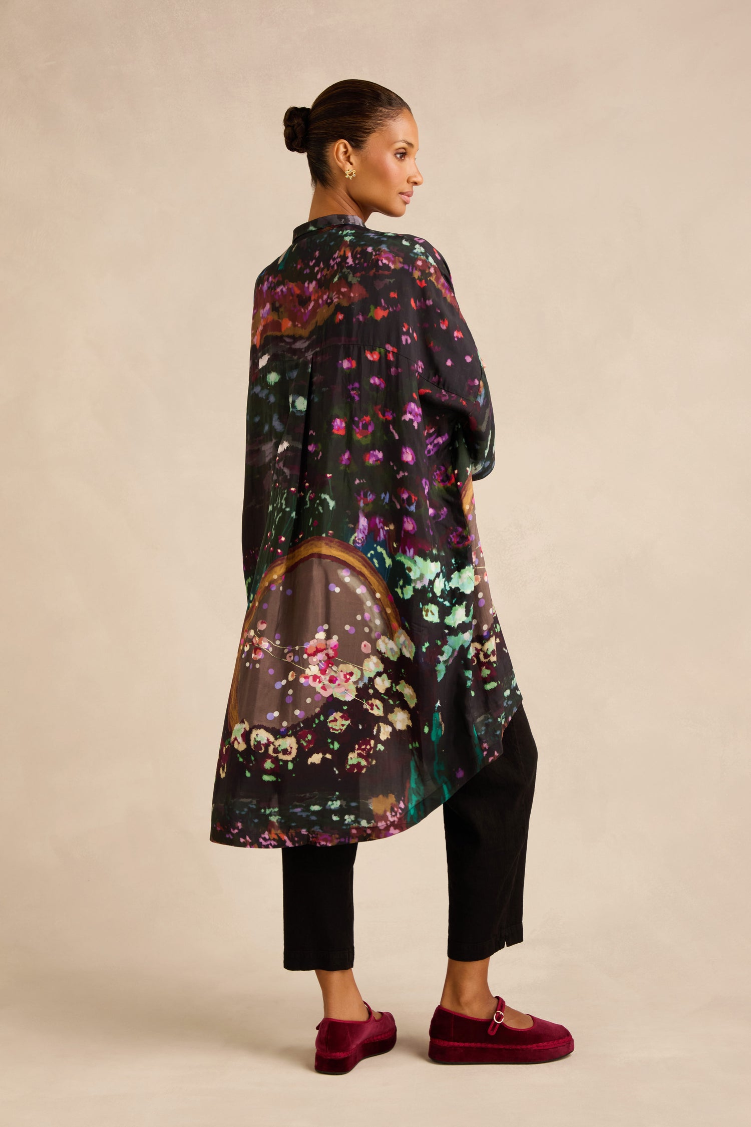 A woman stands sideways against a neutral background, wearing the Water Lilies Button Through Tunic in black with colorful abstract patterns, paired with black pants and red loafers.