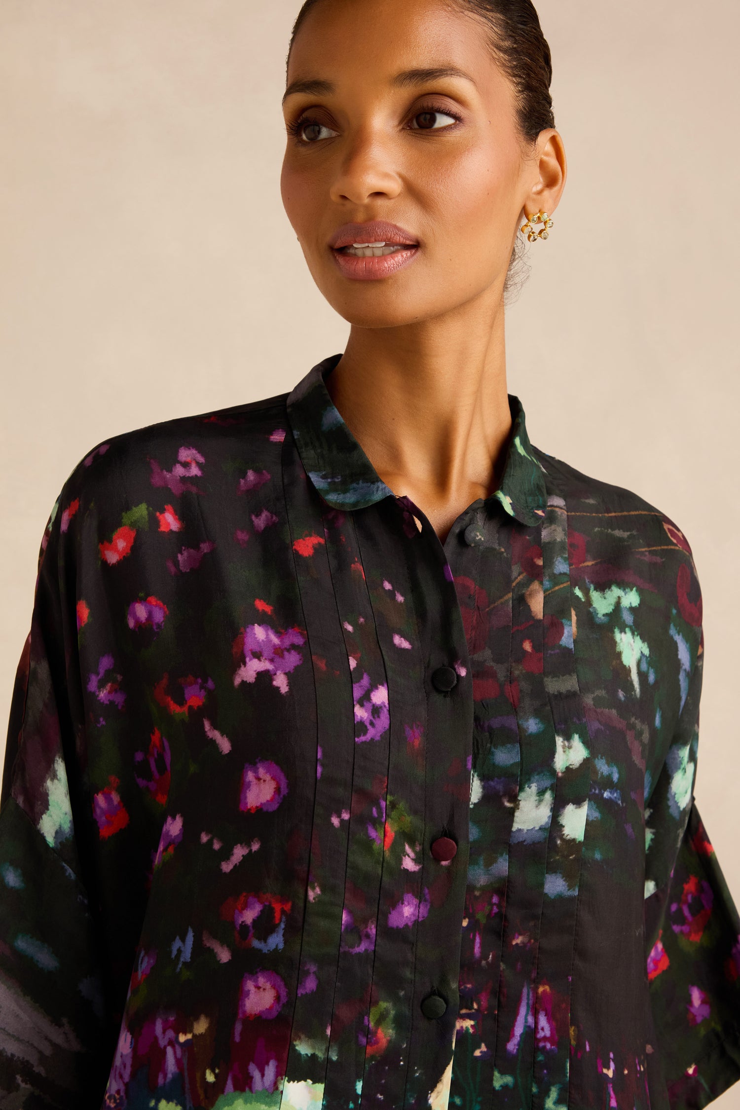 A woman wearing the Water Lilies Button Through Tunic—a black button-up shirt featuring a colorful, abstract floral pattern—looks slightly to her left.