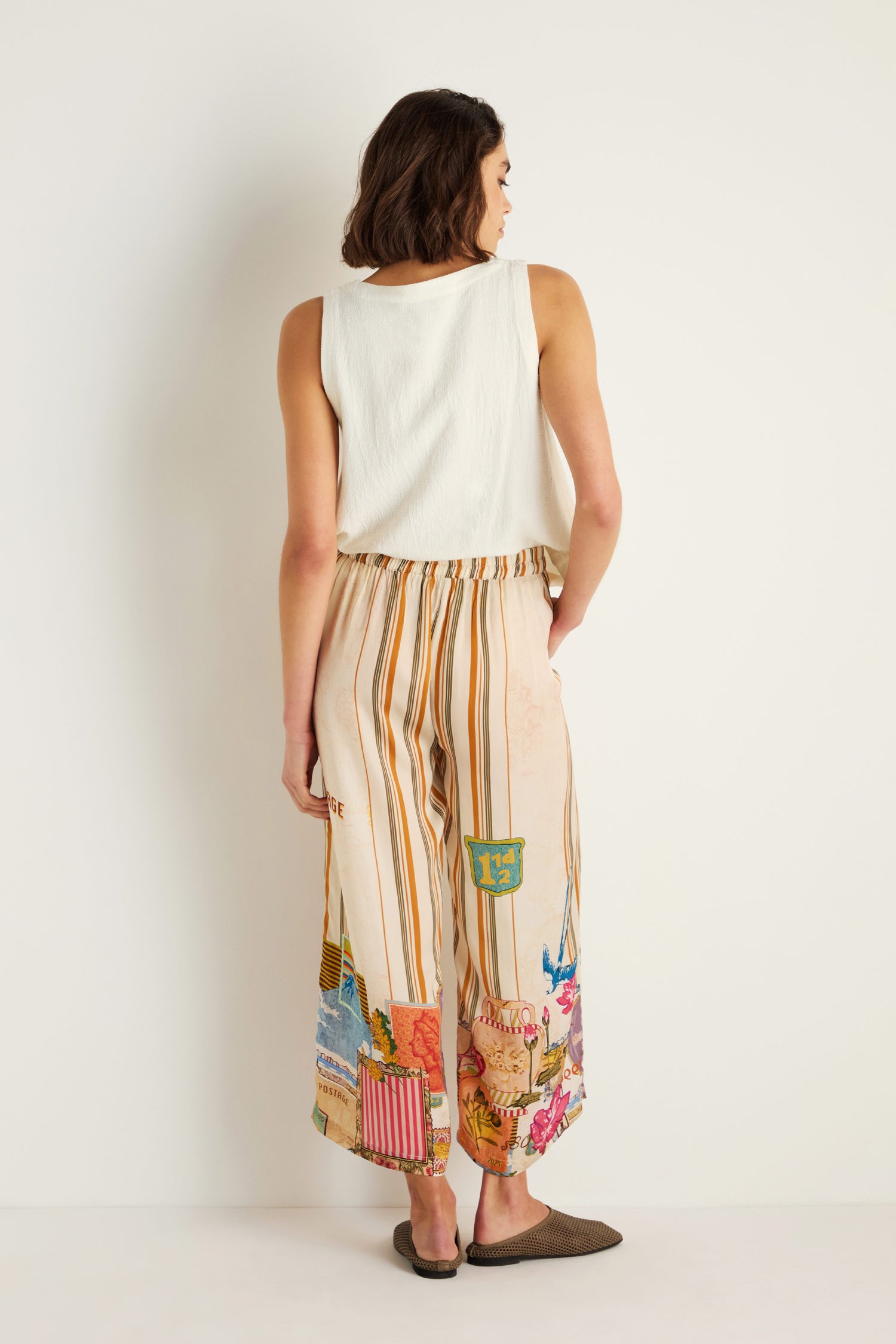 A woman faces away in a sleeveless white top and Postcard Print Stripe Trousers, featuring wide legs and colorful patchwork inspired by Indian textiles, set against a plain light background.