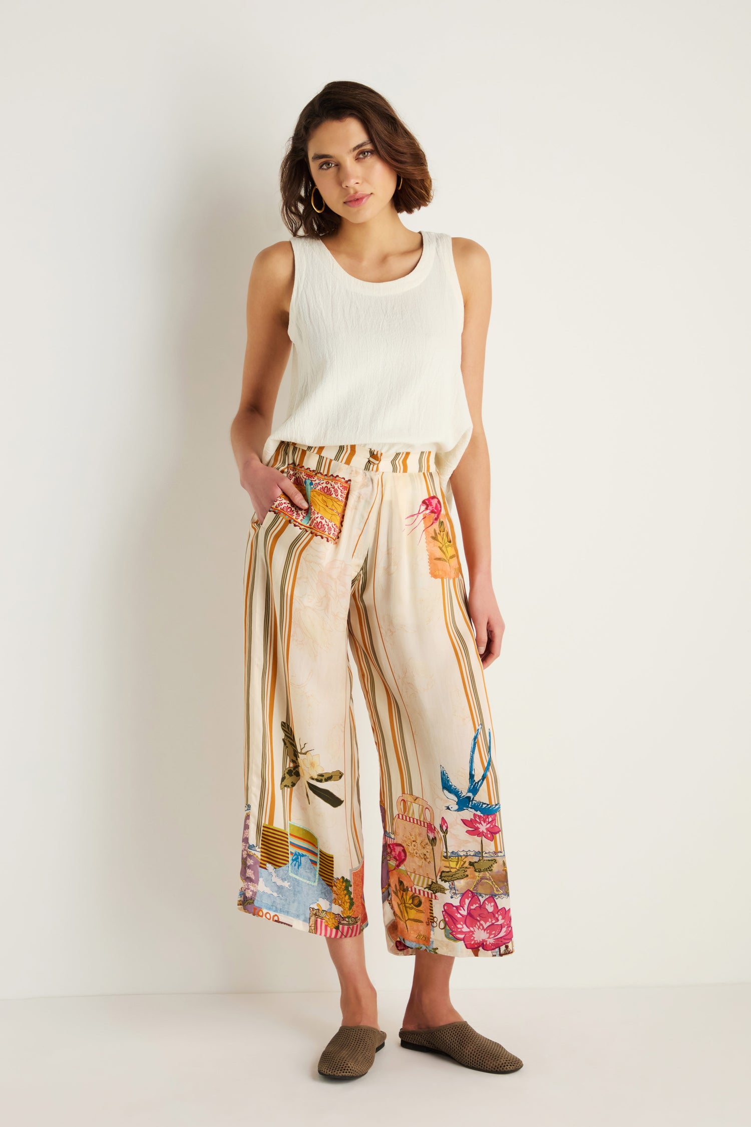 Woman standing against a white wall, wearing a sleeveless white top, Postcard Print Stripe Trousers with colorful floral patterns inspired by Indian textile artistry, and brown slip-on shoes.