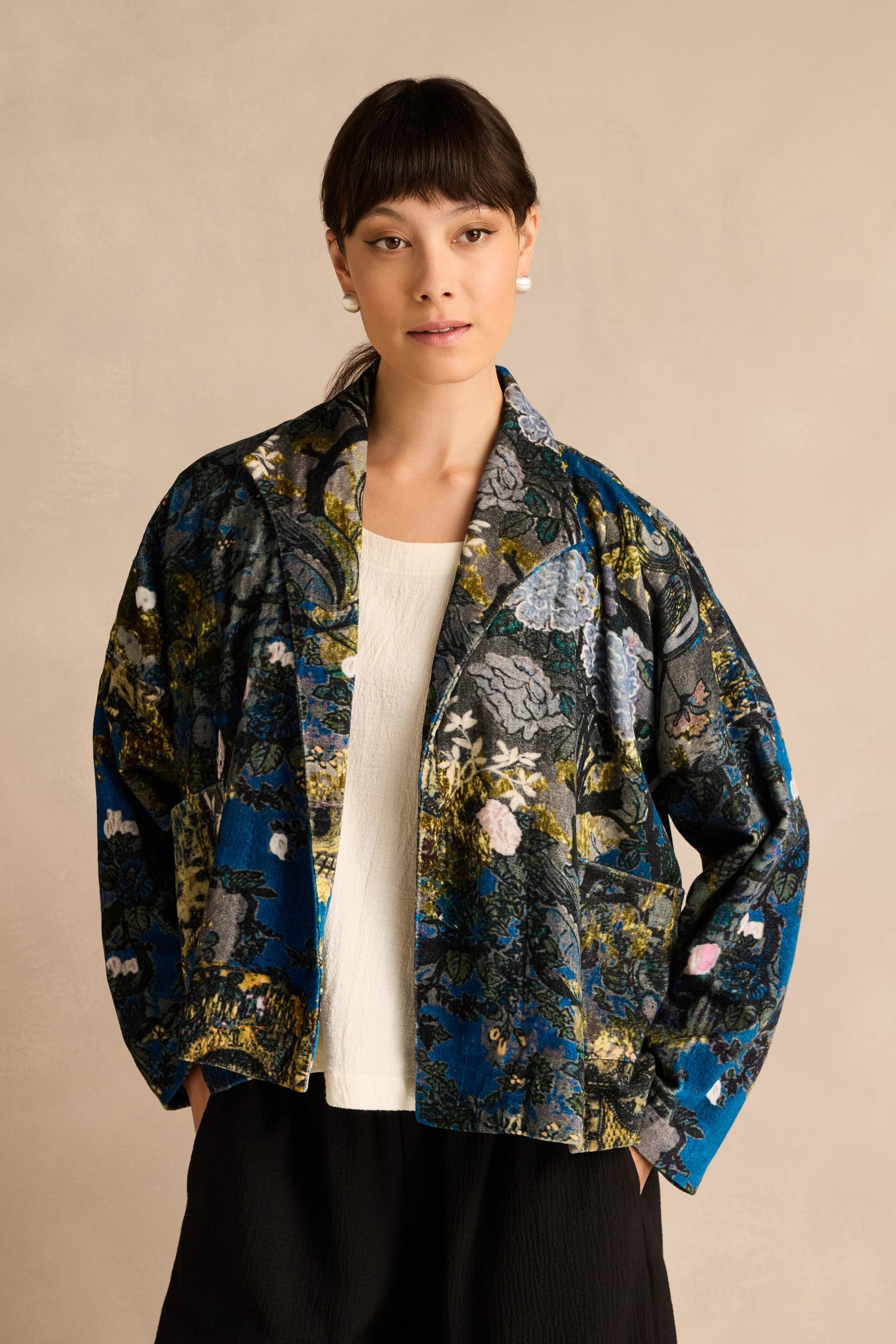 A woman models the Japanese Garden Velvet Jacket, featuring a floral pattern and crafted from artisan-produced cotton velvet, styled over a white top and black pants against a plain beige background.