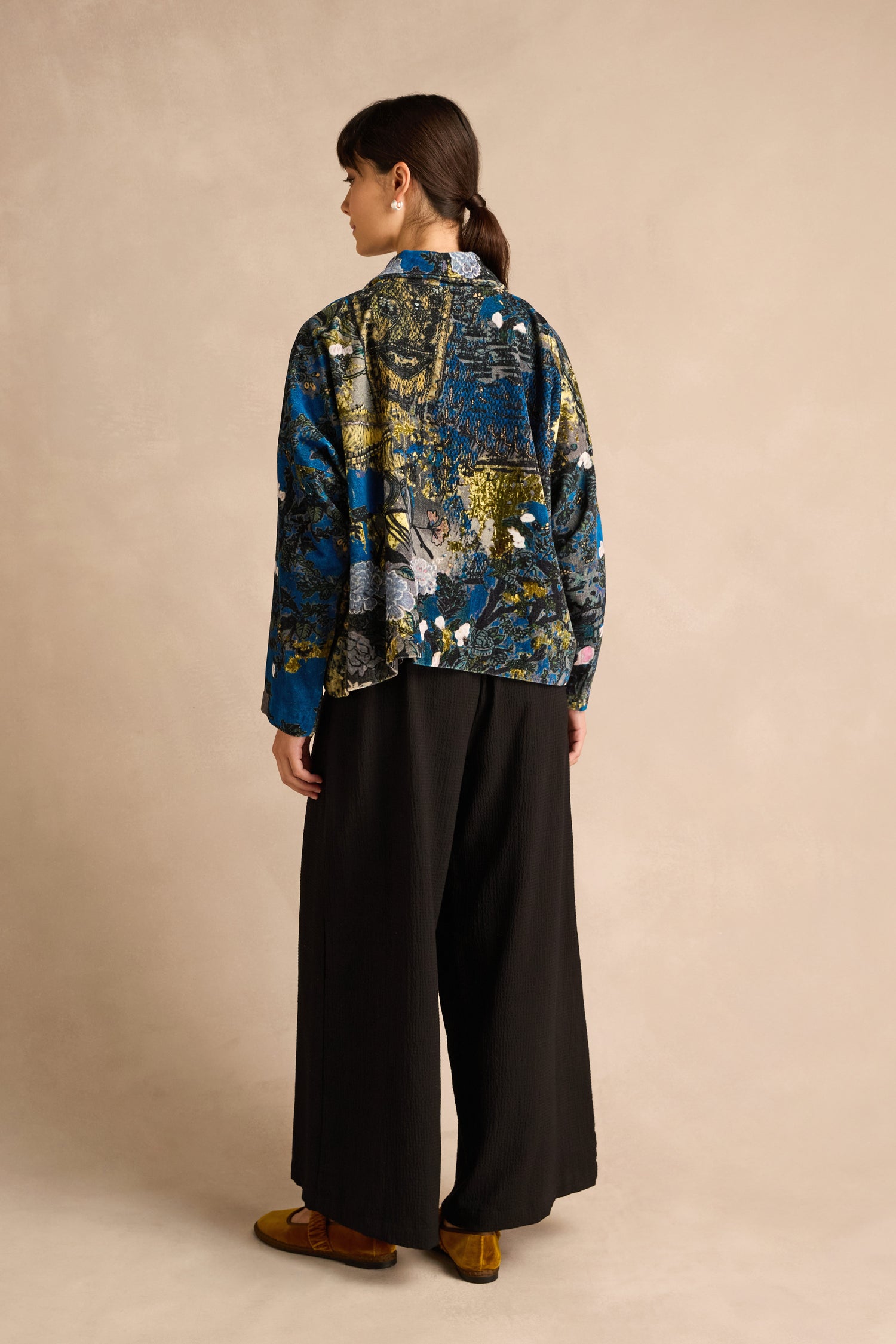 A person stands facing away against a beige background, wearing the Japanese Garden Velvet Jacket—crafted through artisan production in blue and gold—paired with wide black pants and brown shoes.