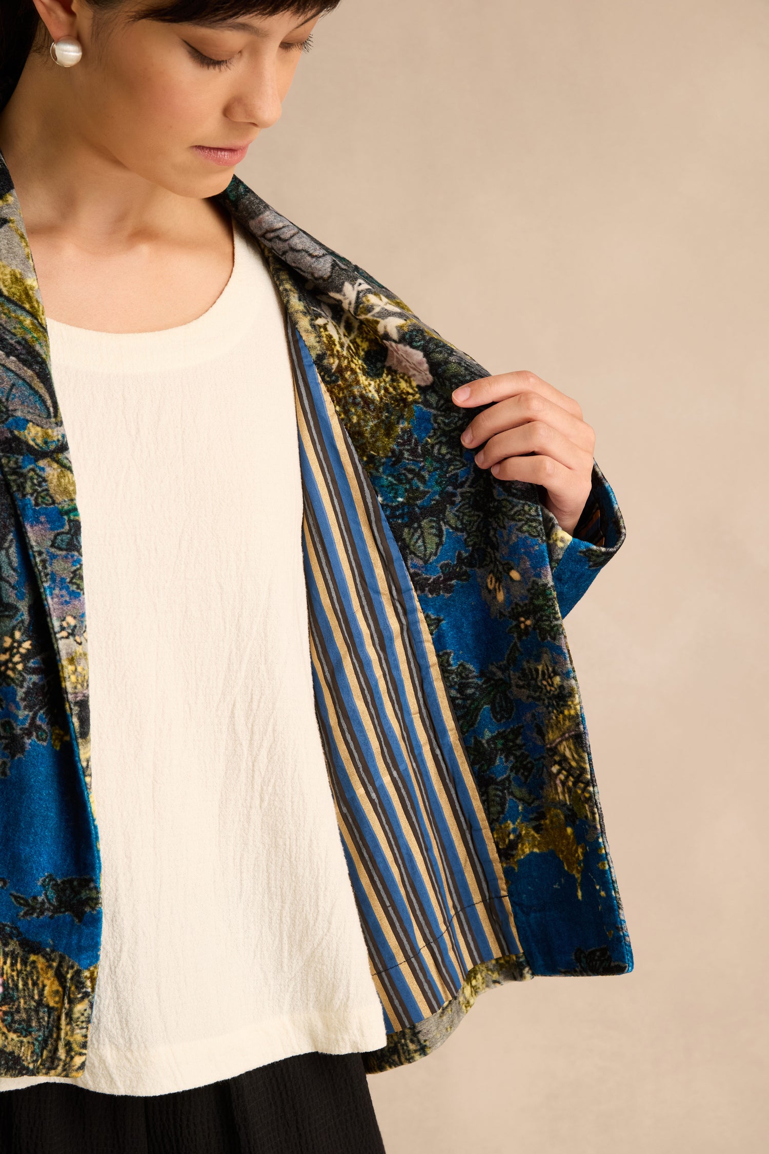 A person opens the Japanese Garden Velvet Jacket to reveal its blue and yellow striped inner lining, styled over a white top. Crafted from cotton velvet, this jacket showcases artisan production with meticulous attention to detail.