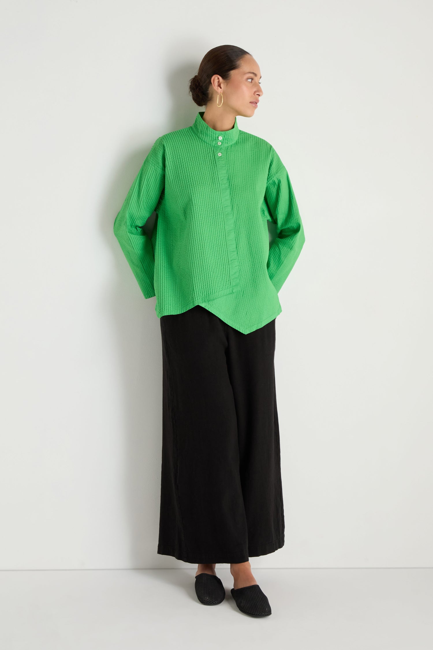 A person stands against a plain white wall wearing an Asymmetric Cotton Bright Green Shirt, wide black pants, black shoes, and large hoop earrings.