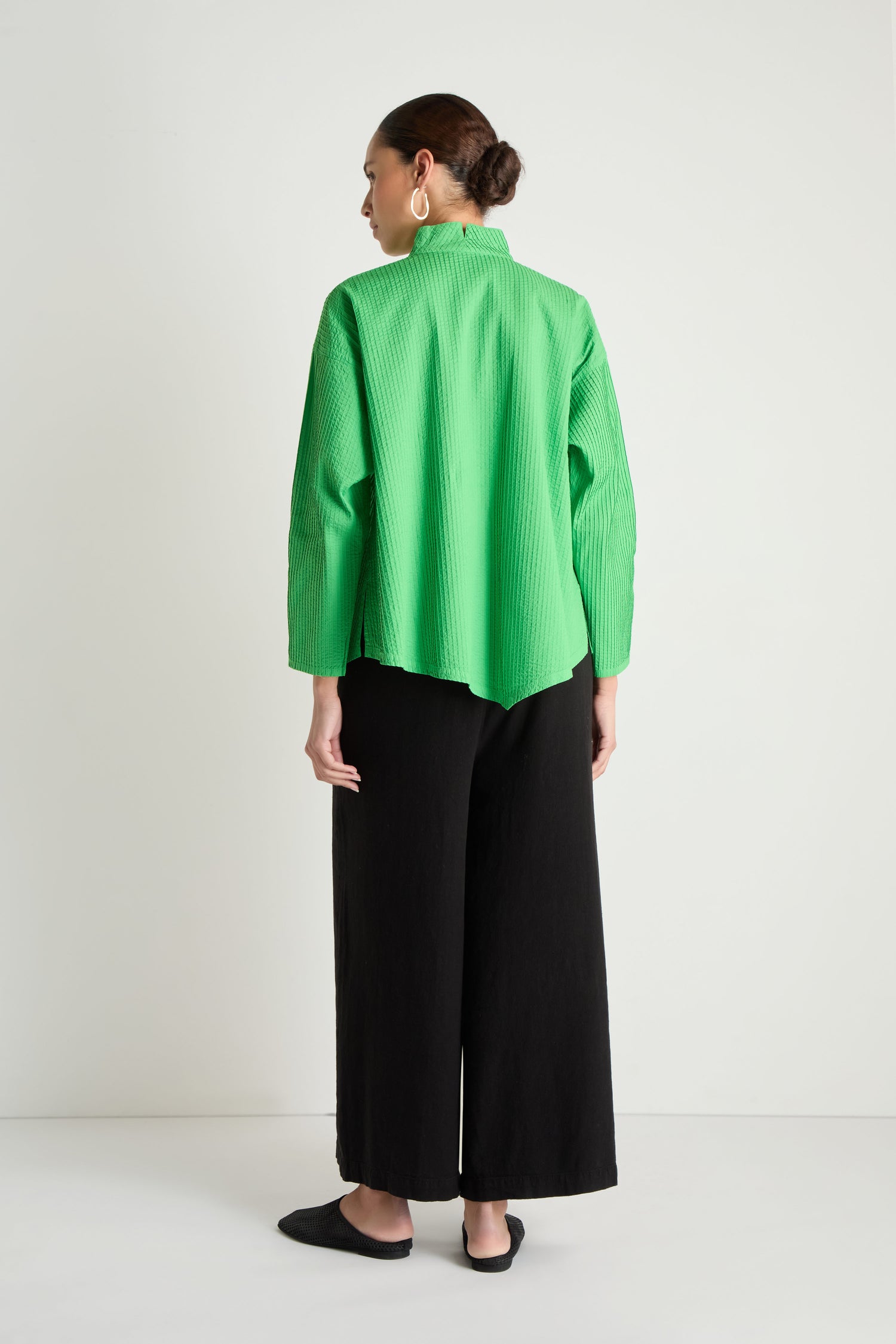 A woman stands facing away, wearing the Asymmetric Cotton Bright Green Shirt with a stand neck, paired with wide black pants, black shoes, and gold hoop earrings. Her hair is tied back in a bun.