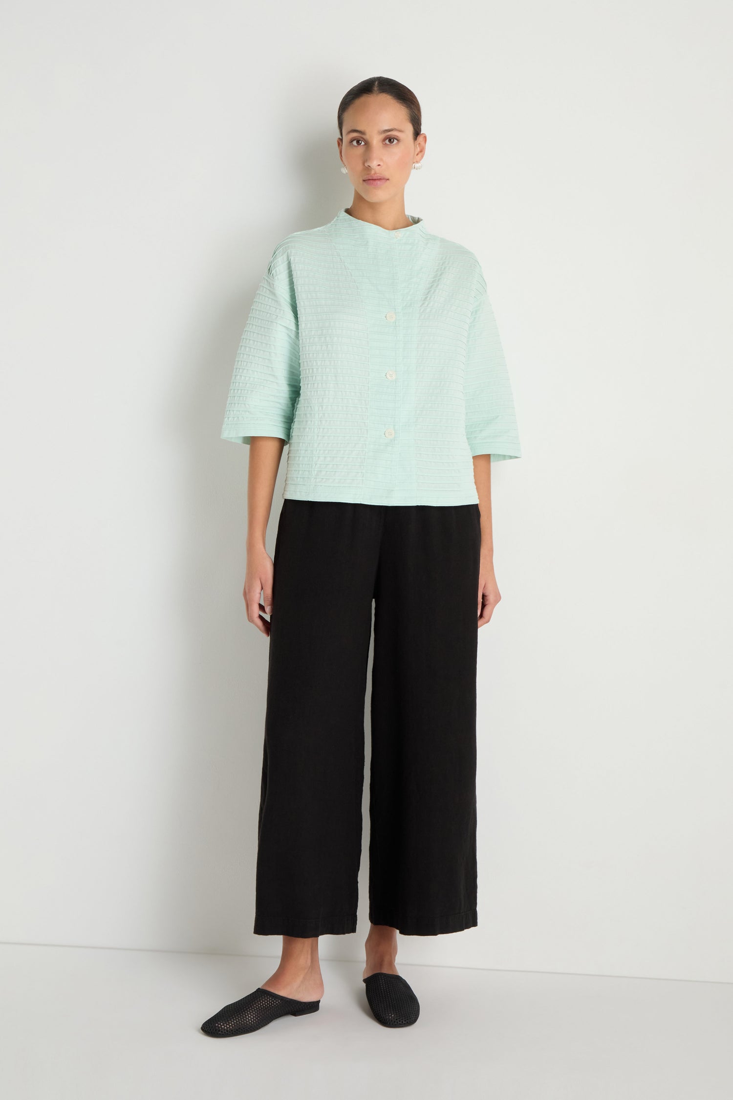 A person stands against a white wall wearing the Funnel Neck Stone Blue Cotton Jacket, black wide-leg pants, and black slip-on shoes.