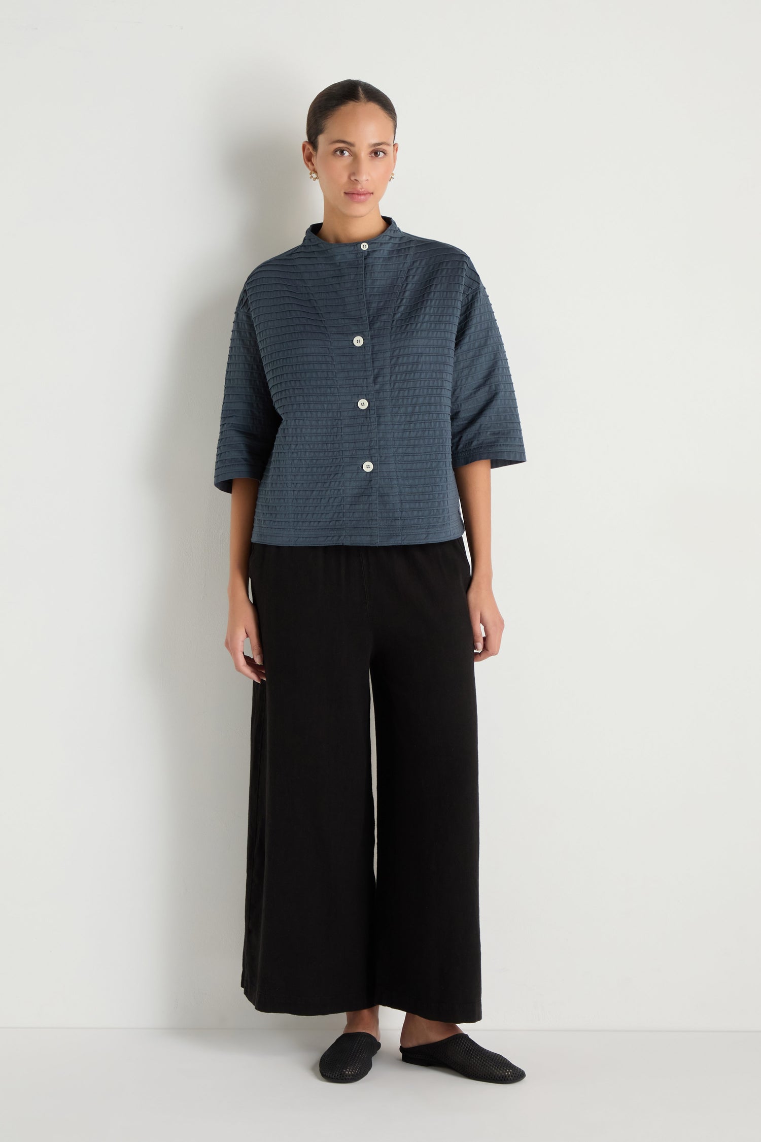 A person stands against a plain white wall wearing the Funnel Neck Stone Blue Cotton Jacket with a boxy silhouette, paired with black wide-leg pants and black shoes.