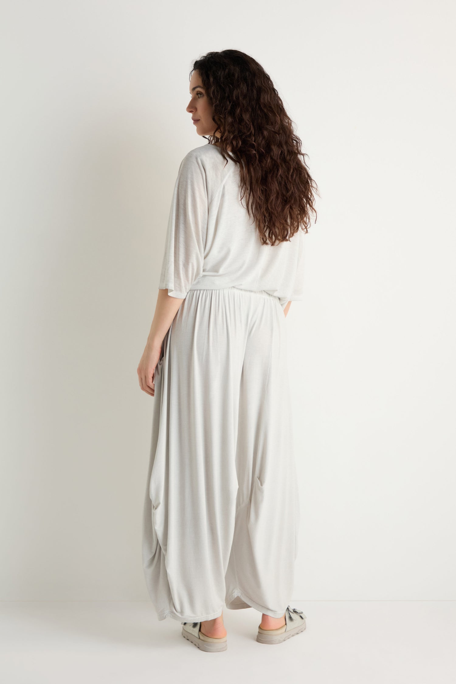 A woman with long curly hair, seen from behind, wears Soft Drape Jersey Trousers and a loose light gray top with casual shoes, standing against a plain white background.