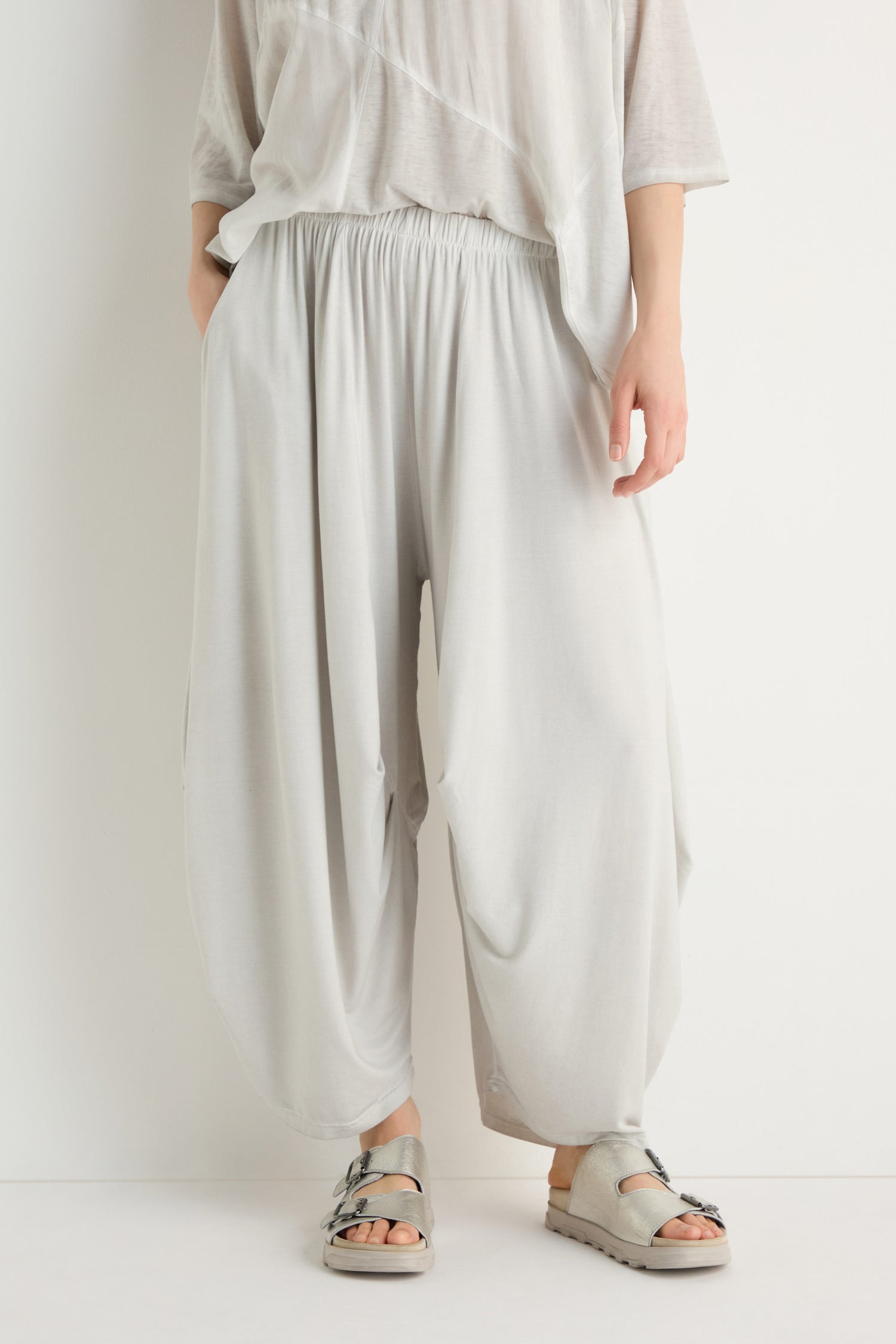 A person stands against a plain white background wearing loose, light gray Soft Drape Jersey Trousers, a matching top, and gray sandals.
