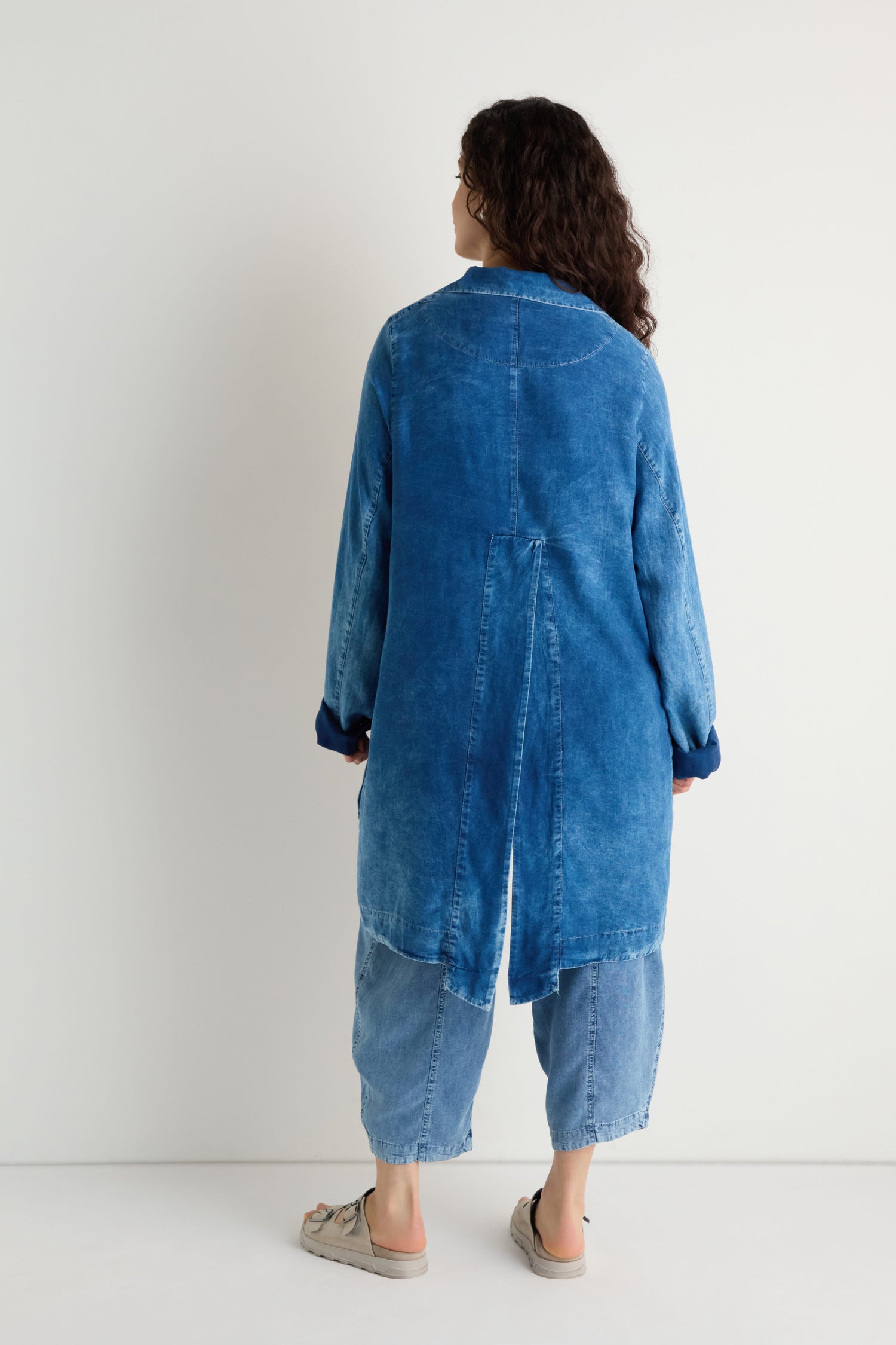 A person stands facing a white wall, wearing a Lyocell Utility Jacket in washed blue with a relaxed fit, paired with loose-fitting jeans and beige shoes.