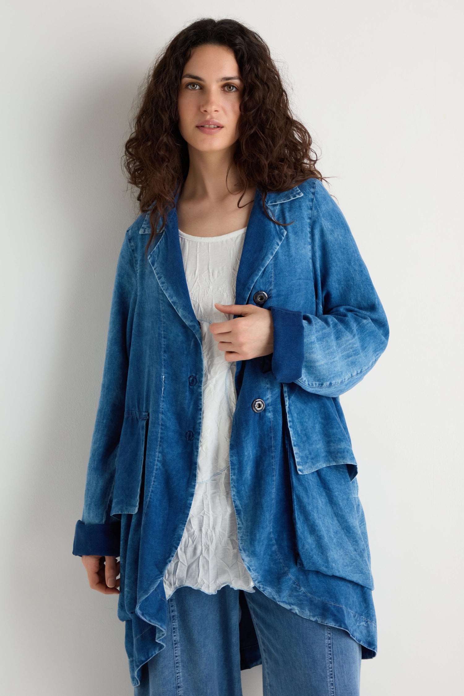 A person with curly brown hair wears a loose Lyocell Utility Jacket in blue over a white textured top and blue jeans, standing against a plain light-colored wall.