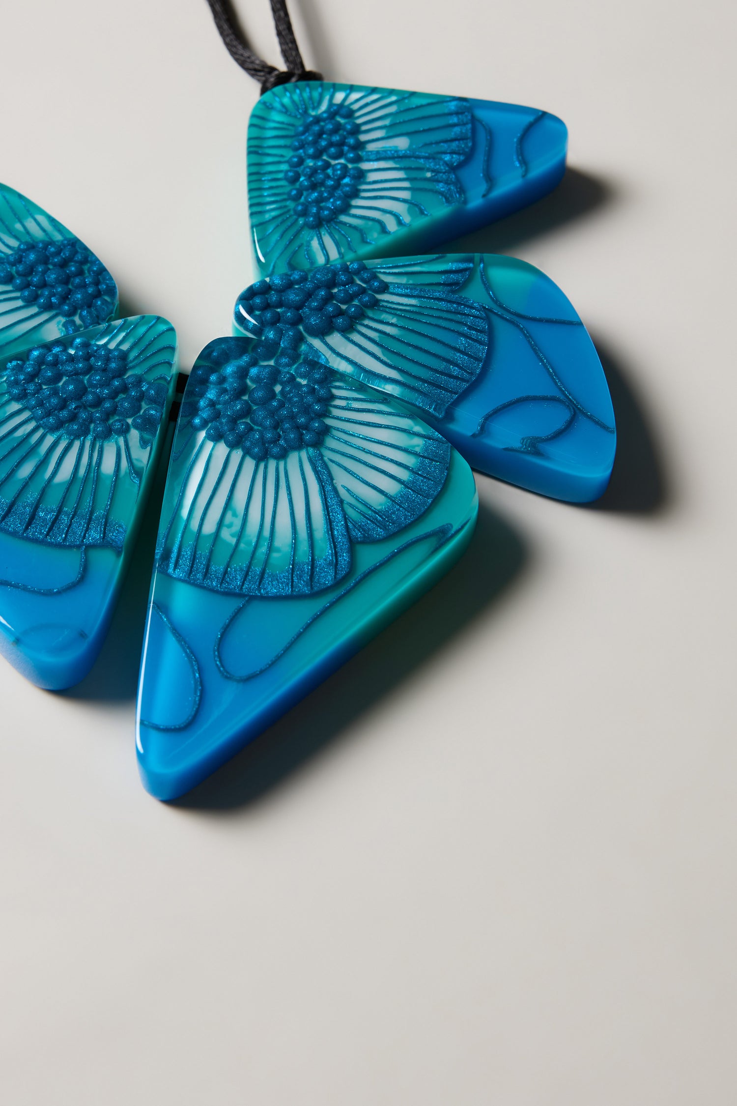 Close-up of the Primula Resin Necklace, featuring three blue triangular pendants with delicate patterns in Zsiska style, displayed on a light surface.