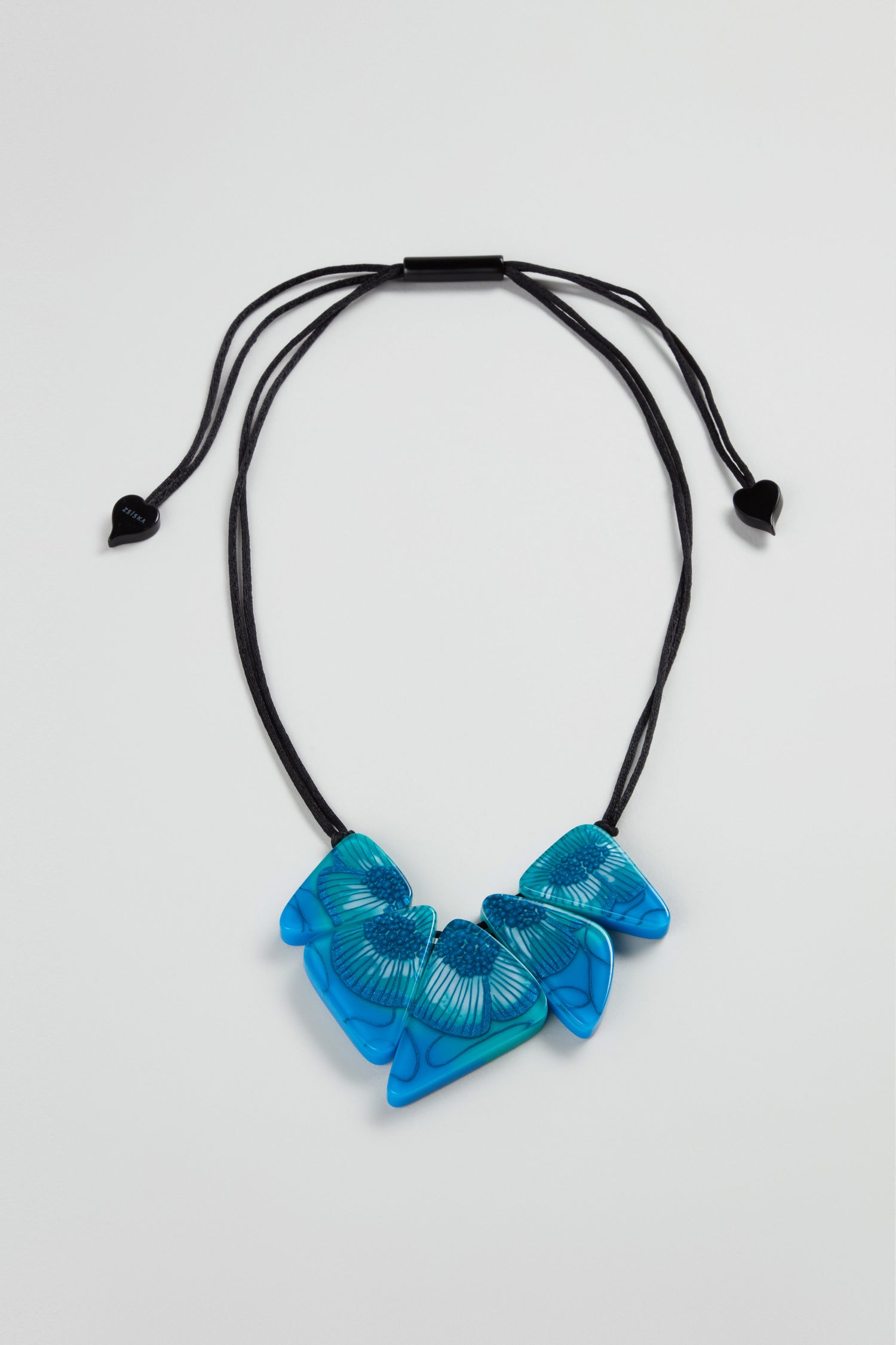 The Primula Resin Necklace by Zsiska, with three blue wing-shaped pendants on a black adjustable cord, is displayed against a light gray background—a striking piece of modern jewellery.