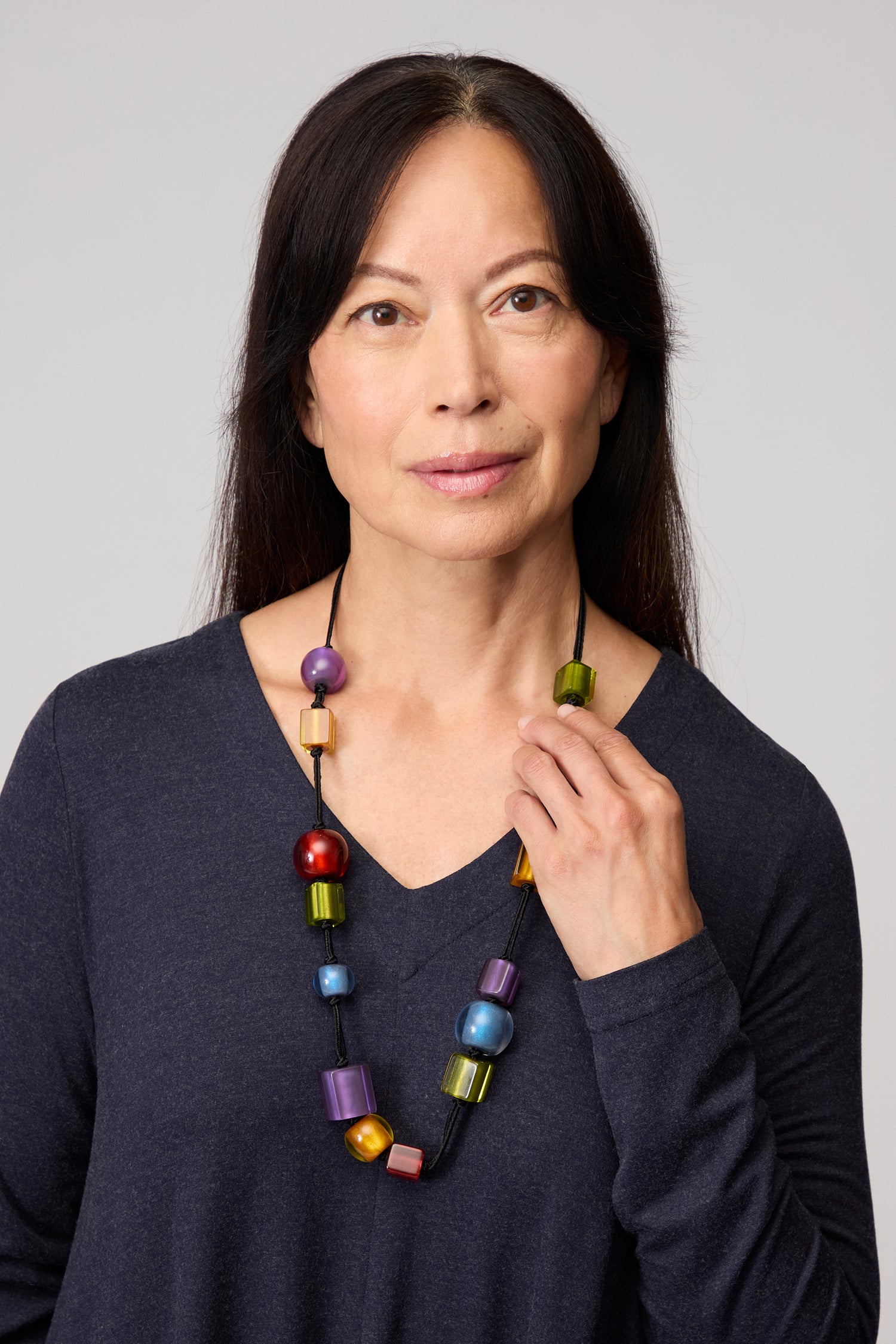 A woman with long dark hair, dressed in a dark V-neck top, showcases the Colourful Shapes Resin Necklace while standing against a plain light background.
