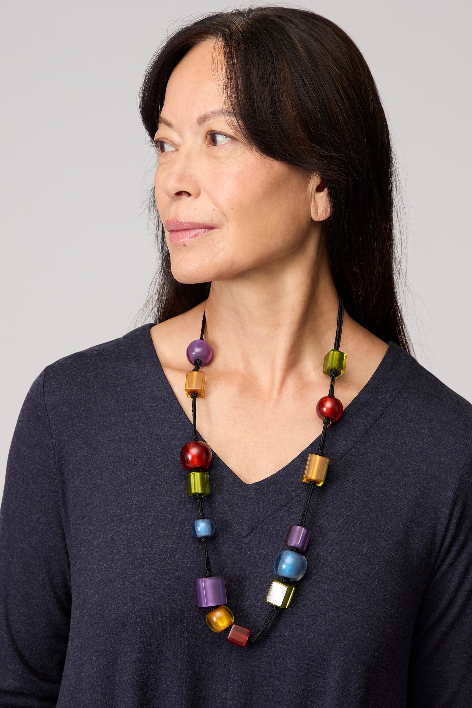 A woman with long dark hair, dressed in a navy V-neck top, gazes left against a plain backdrop while wearing the Colourful Shapes Resin Necklace—a vibrant handmade piece that adds a bold pop of color to her look.