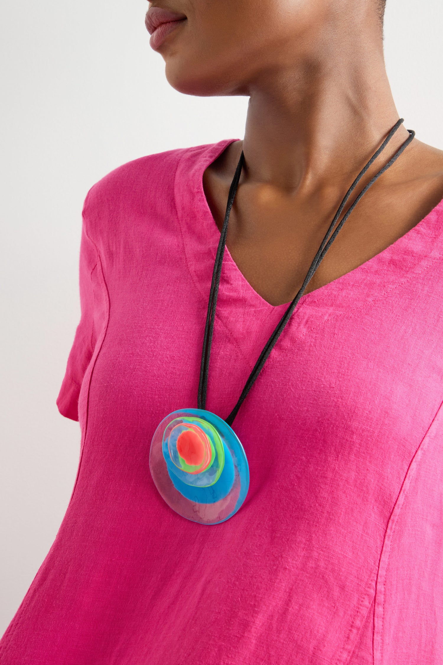 A person is wearing a pink top and the Blue Neon Flora Pendant, featuring a vibrant circular design. This handcrafted resin piece elegantly hangs on an adjustable cord.