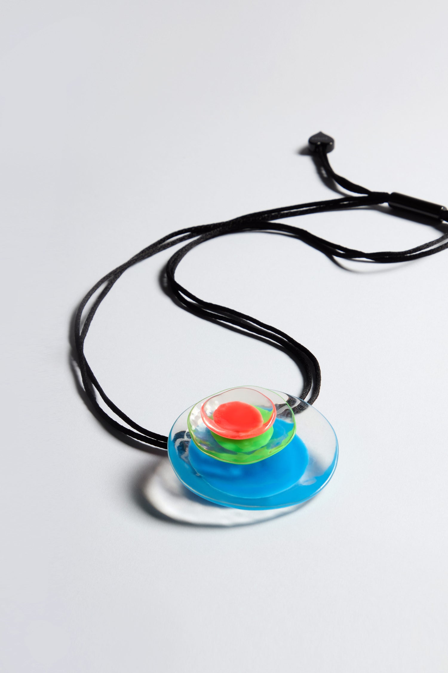 The Blue Neon Flora Pendant from the FLORA collection showcases a handcrafted resin design with concentric layers of blue, green, and red colors on an adjustable black cord.