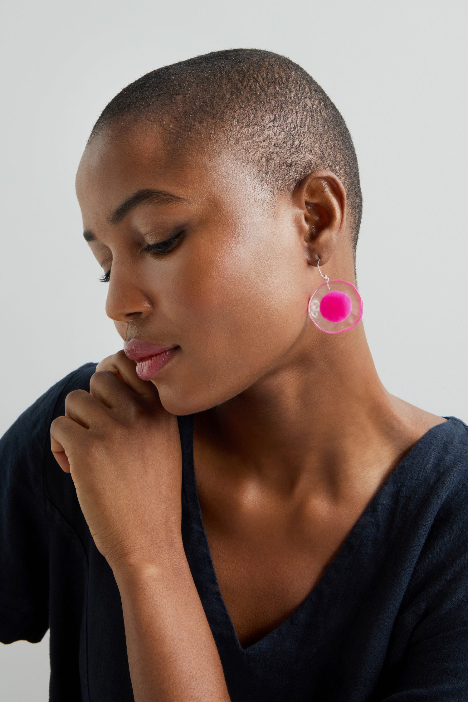 A person with short hair in a black top thoughtfully gazes downward, wearing vibrant pink Flora Earrings against a plain backdrop.