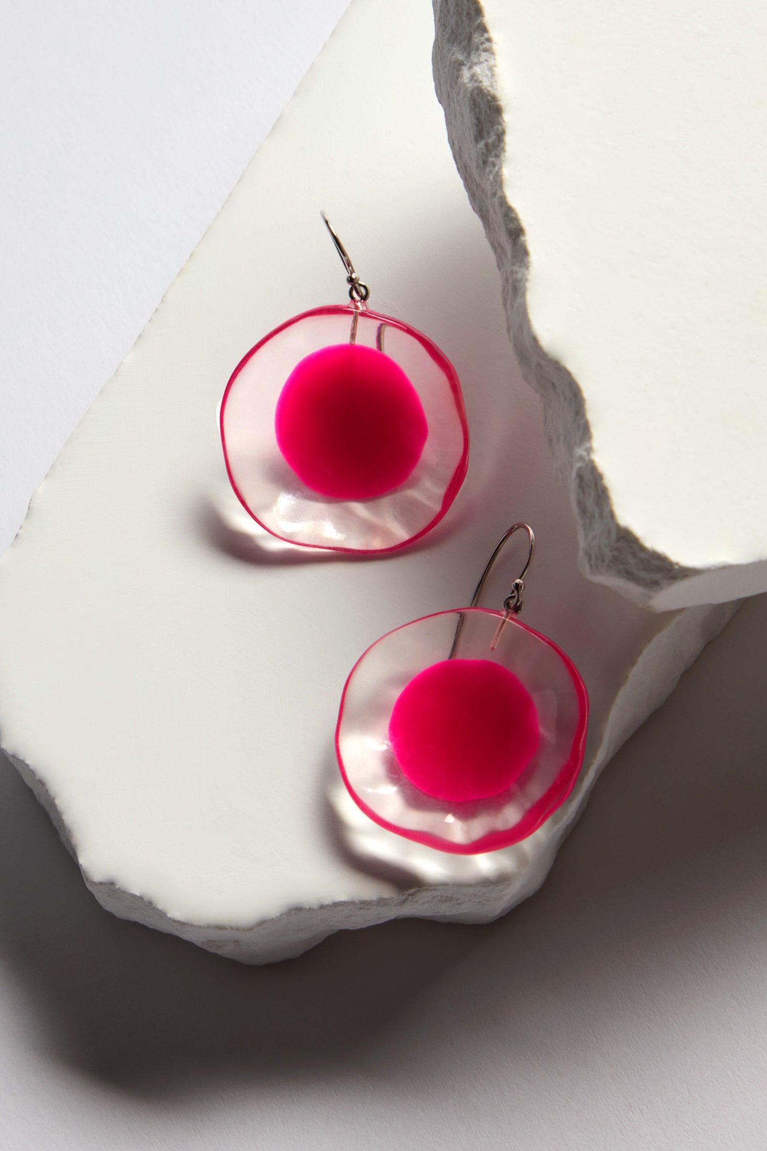 The Flora Earrings, part of the Prue by Zsiska collection, display artistry with handcrafted resin featuring clear circular discs and vibrant pink spheres on white textured surfaces.