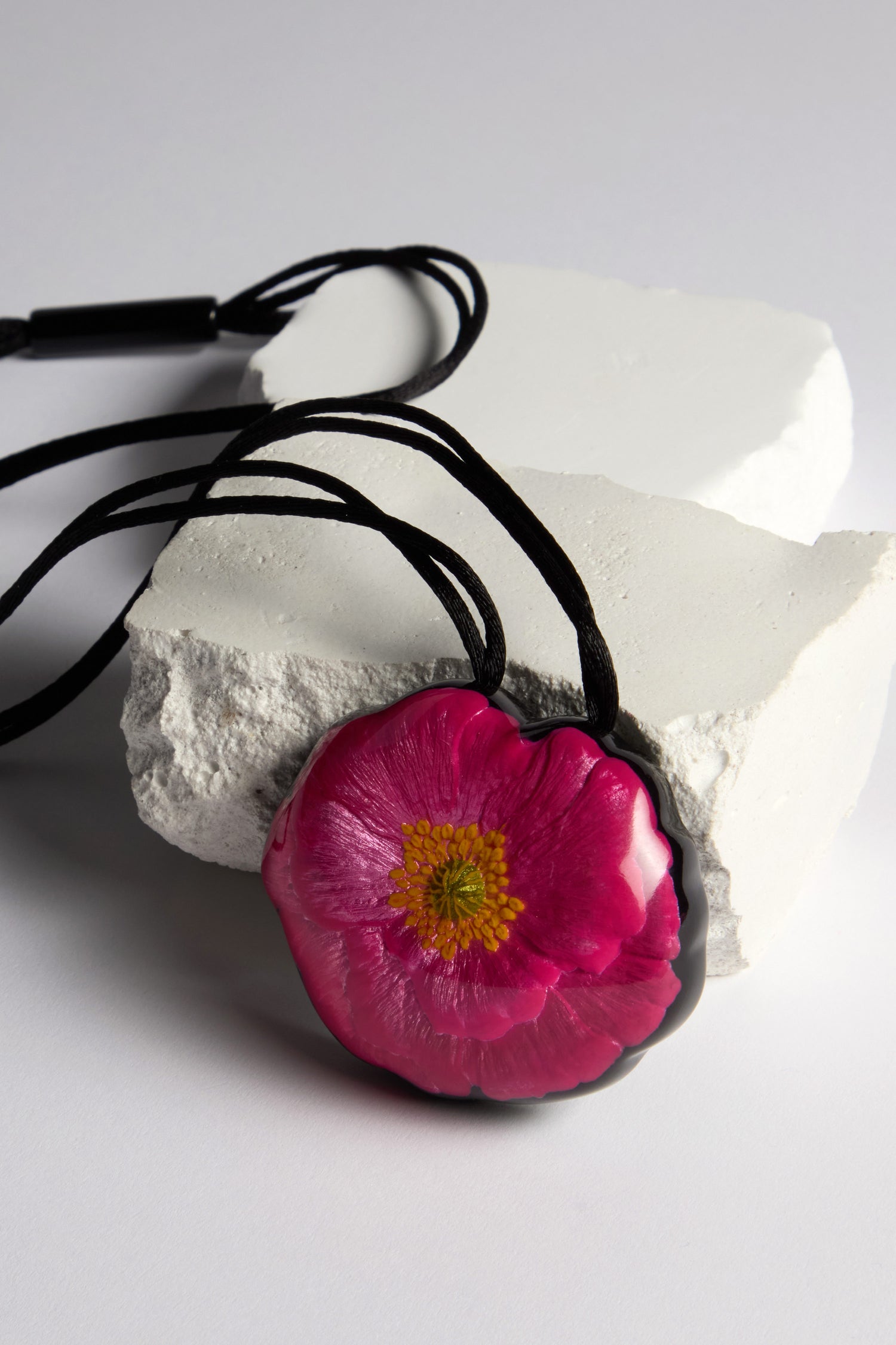 The Garland Pendant, a handcrafted piece showcasing a pink flower with a yellow center, hangs gracefully from an adjustable black cord on a textured white rock.