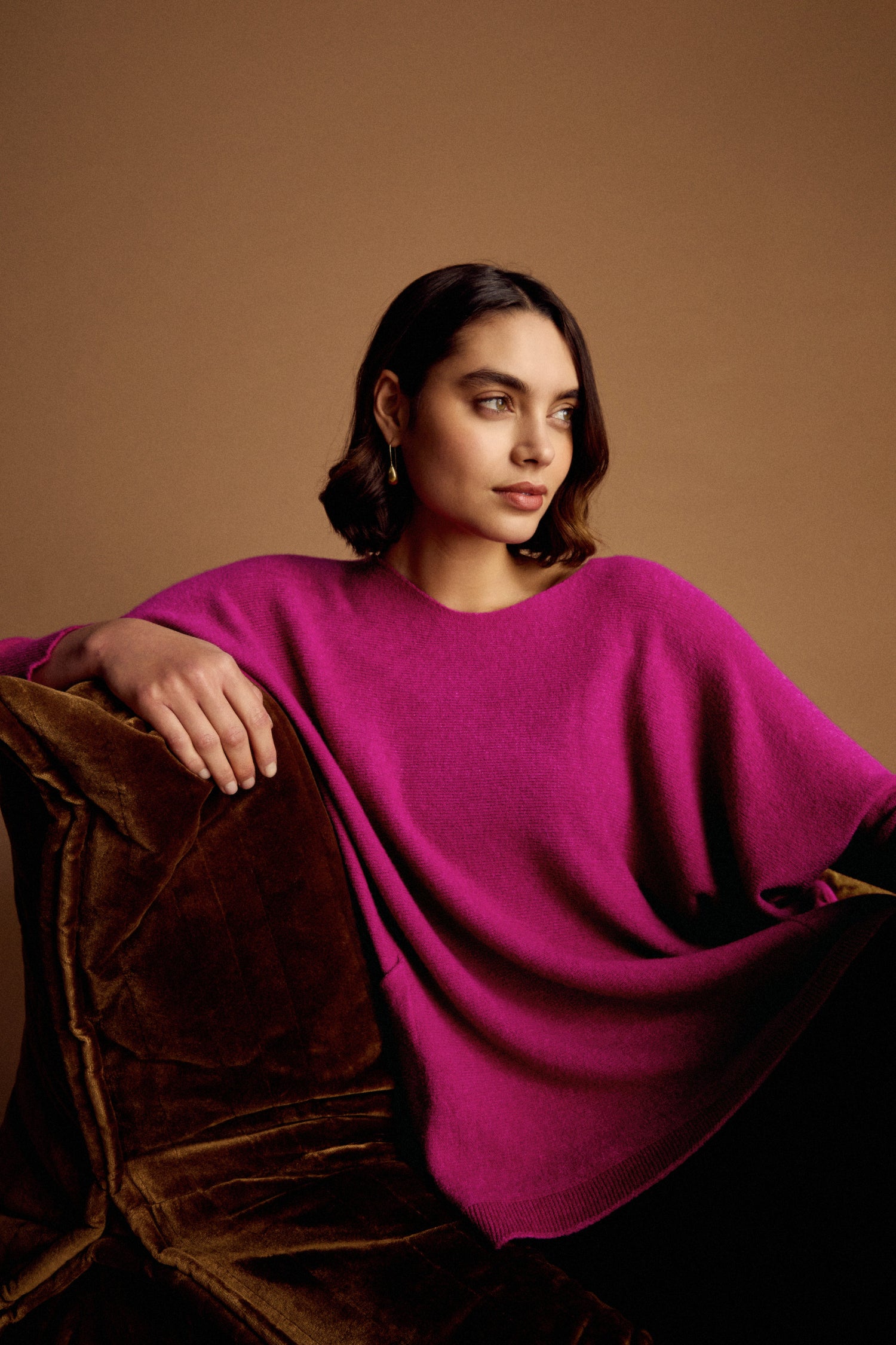 A woman with dark hair and gold earrings sits on a brown velvet chair, wearing the Two Pocket Oversize Knit in bright magenta, against a plain brown background.