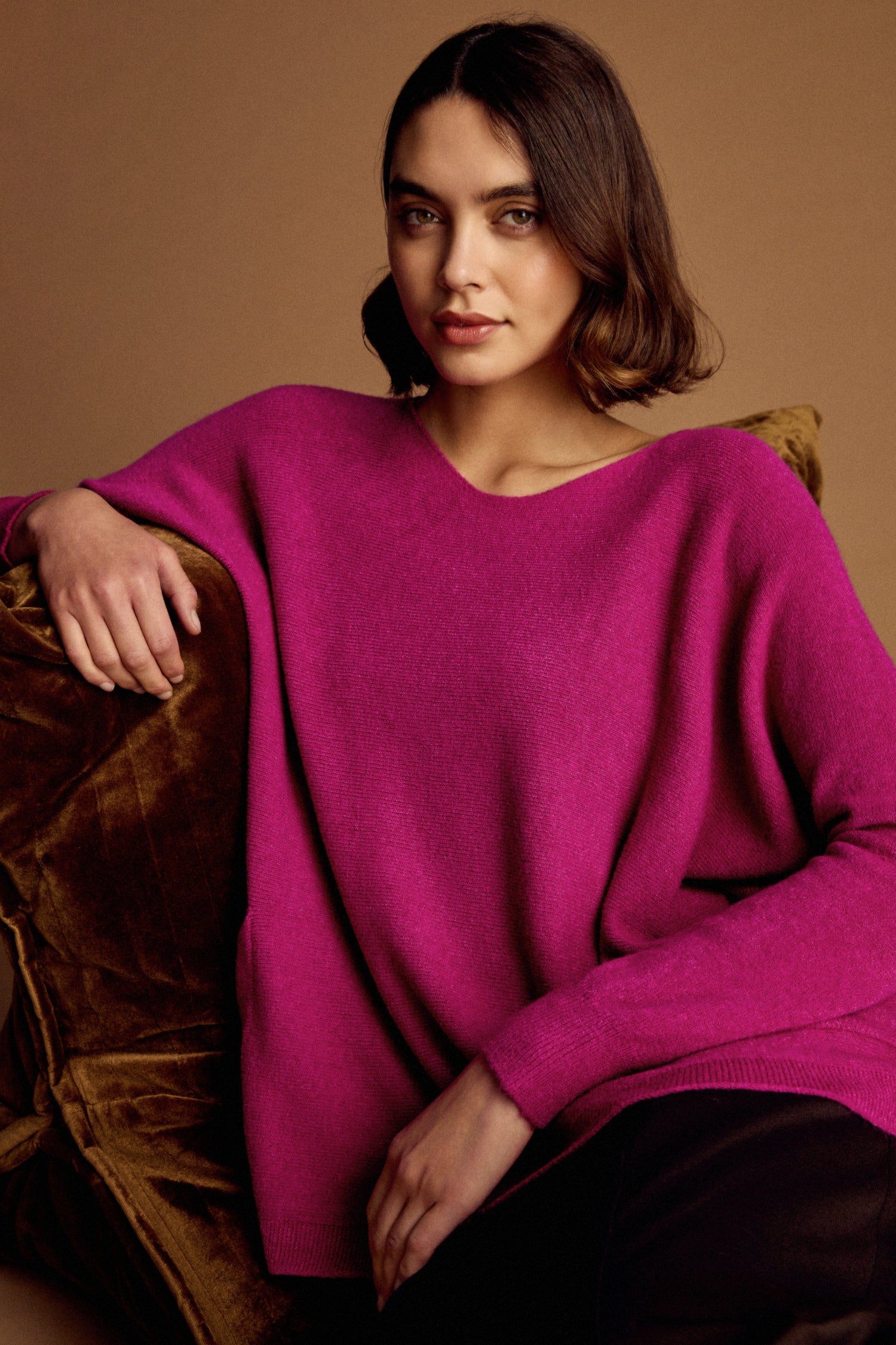 A woman with short brown hair wears the bright pink Two Pocket Oversize Knit as she sits on a brown velvet chair against a neutral background.