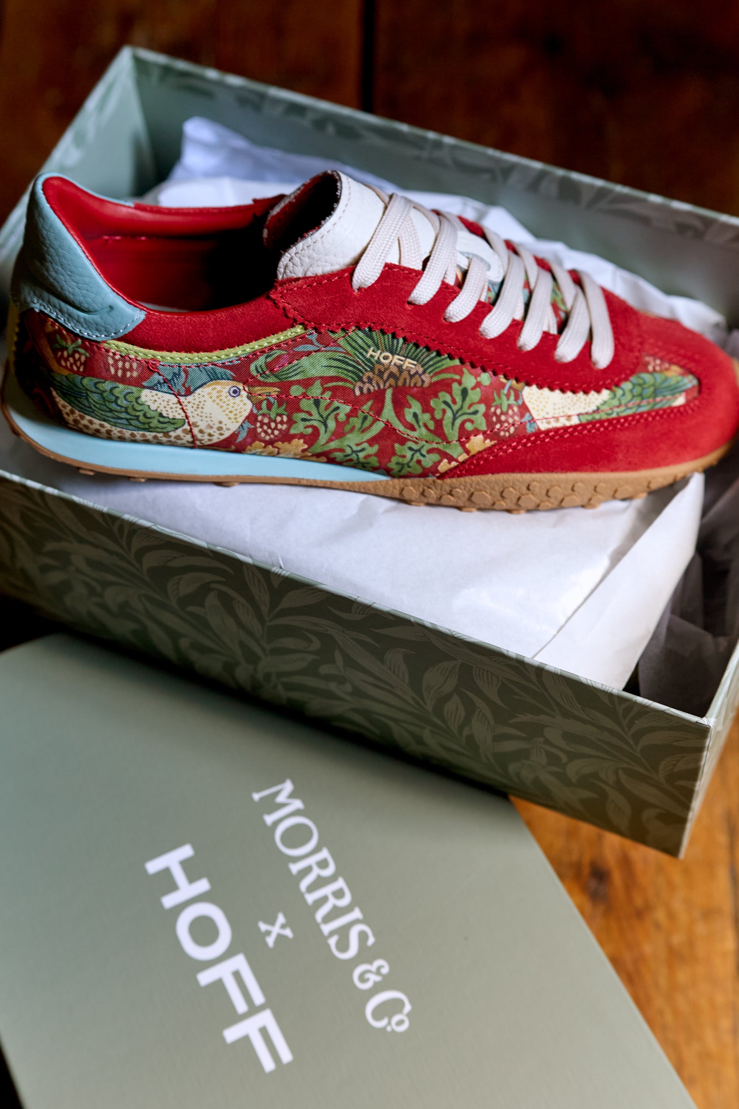 A Red William Morris Bridge Trainer with a red and green pattern sits in an open shoebox featuring “MORRIS & CO. x HOFF” on the lid.