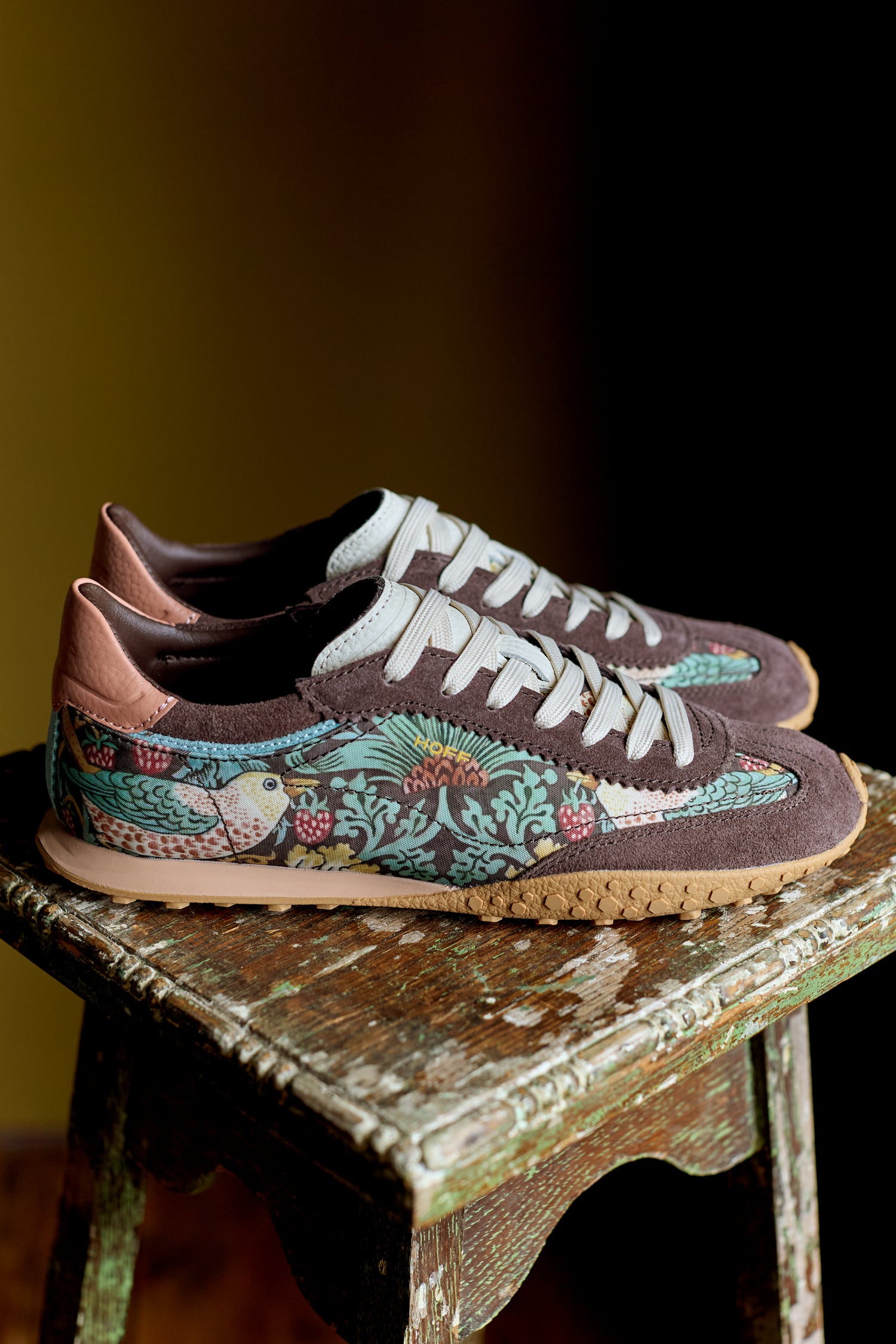 The Brown William Morris Bridge Trainers for women feature white laces and a vibrant bird and floral heritage pattern, displayed on a weathered wooden stool.