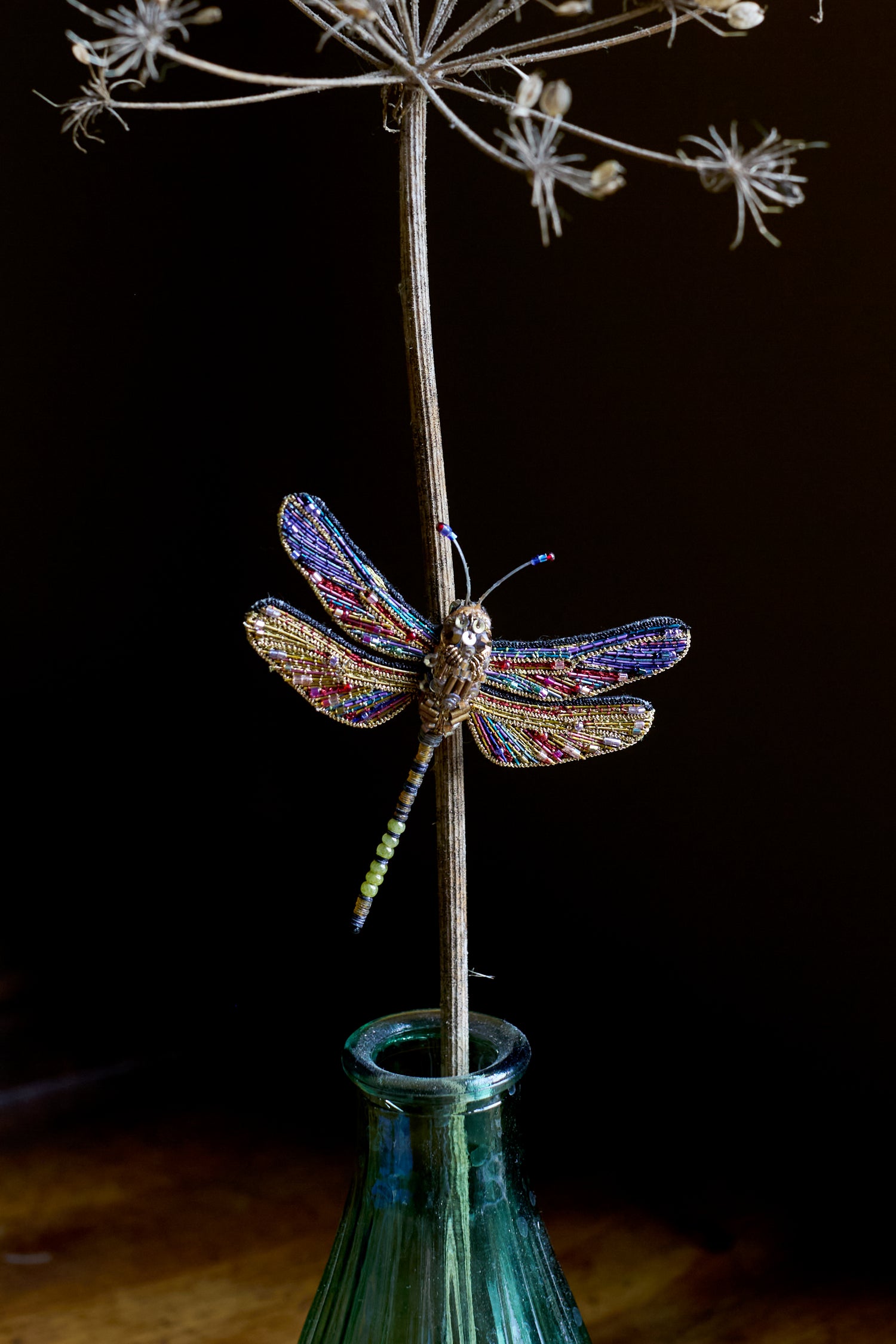 The Hand Beaded Hawker Dragonfly Brooch, a distinctive piece of handmade jewelry, adorns a dried plant stem in a green glass bottle set against a dark backdrop.