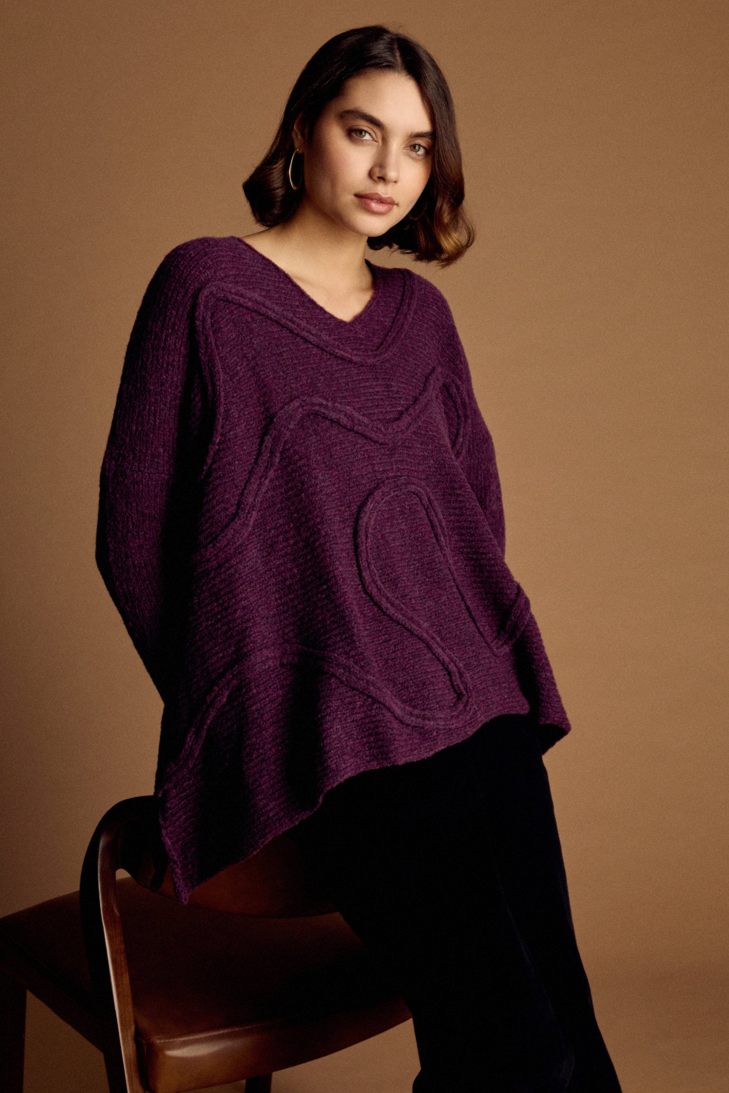 A woman wears the Swirl Seamed Oversize Aubergine Knit sweater with black pants, seated on a brown chair against a plain brown background.