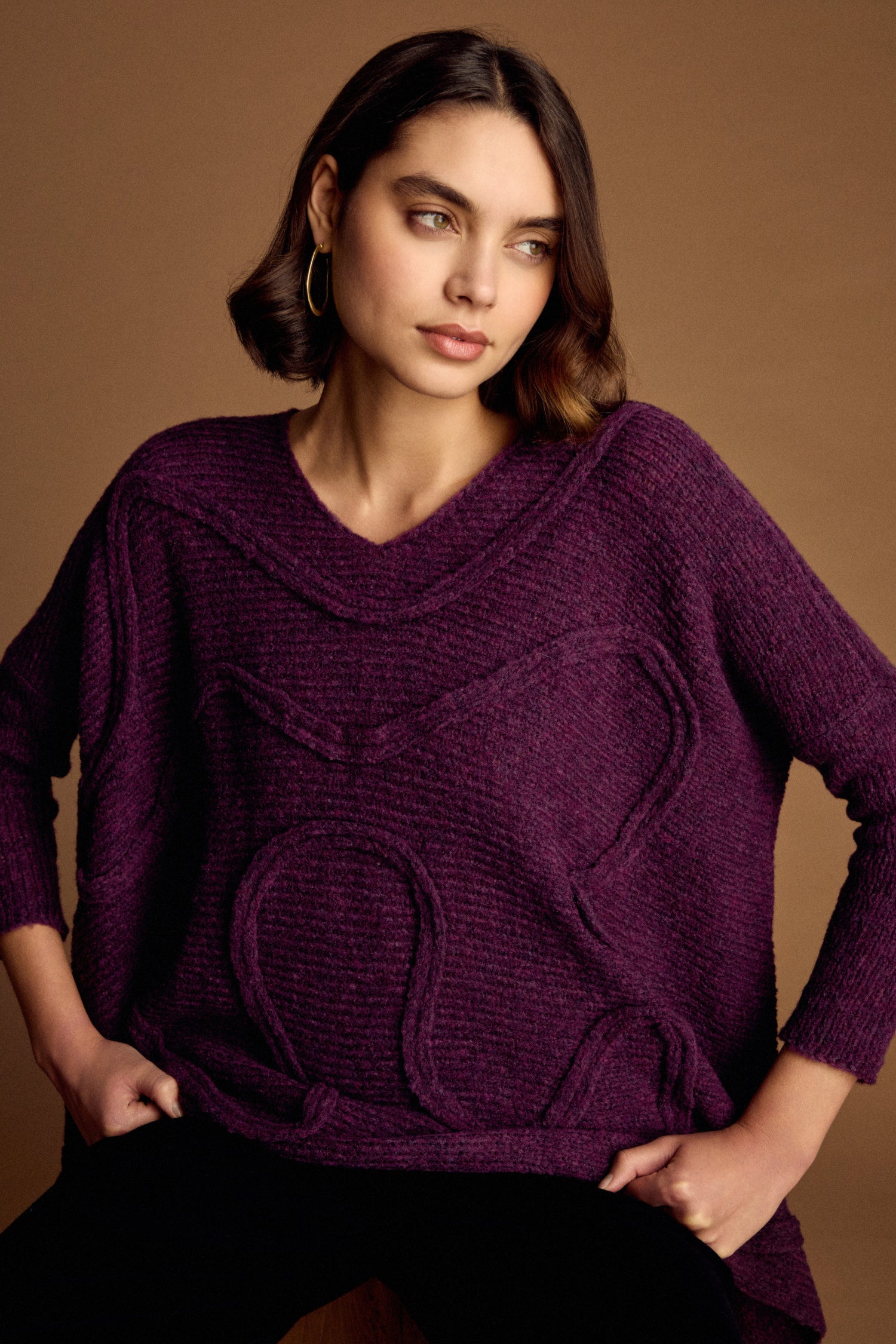A woman with short brown hair wears the Swirl Seamed Oversize Aubergine Knit, sitting against a brown background and gazing to the side with a neutral expression.
