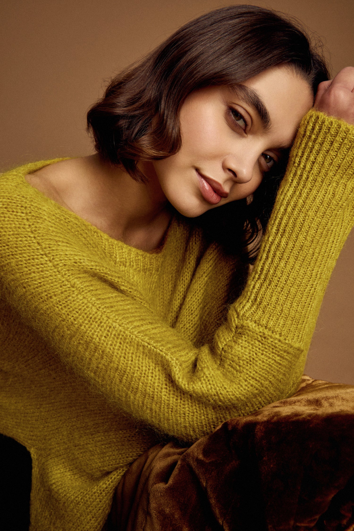 A woman with shoulder-length brown hair wears the Wide Neck Mohair Knit and rests her head on her arm, looking at the camera against a plain brown background.