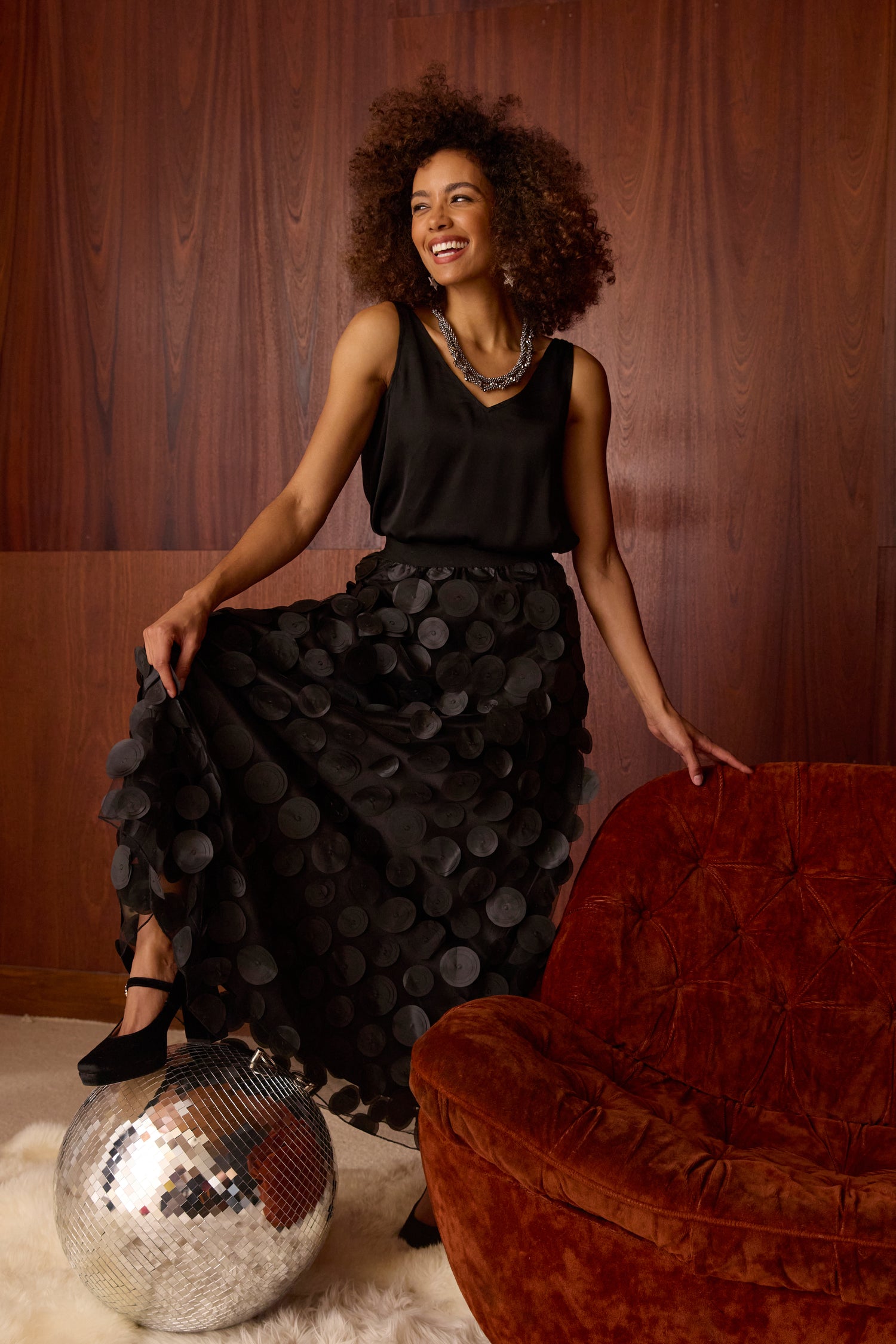 Woman wears a black sleeveless top and the Cut Out Circles Skirt, posing by a brown chair with one foot on a disco ball, set against a wood-paneled wall.