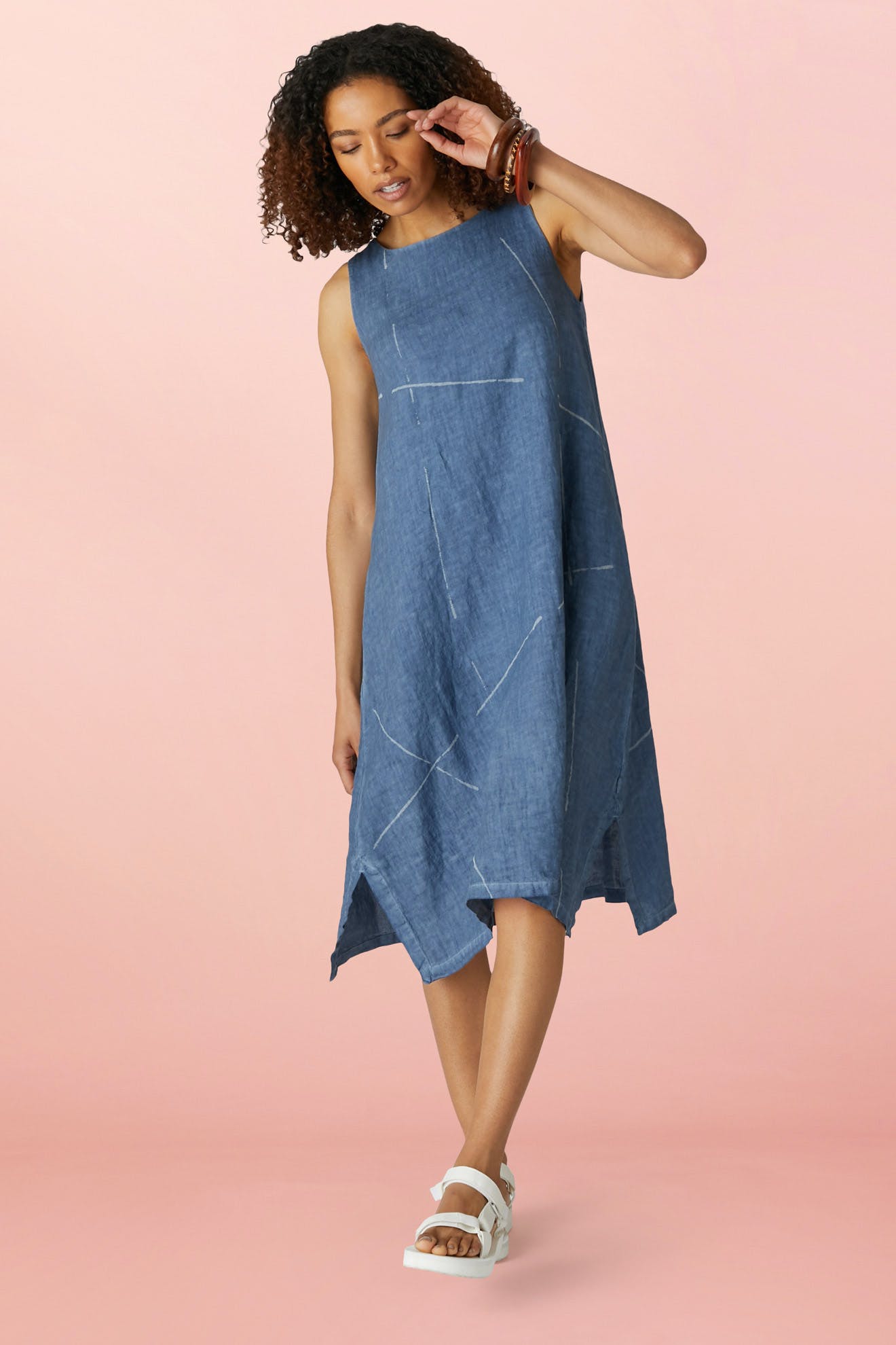 A woman wearing the Som Linen Dress in blue, featuring a relaxed A-line silhouette, and white sandals stands against a light pink background, looking downward with one hand near her face.