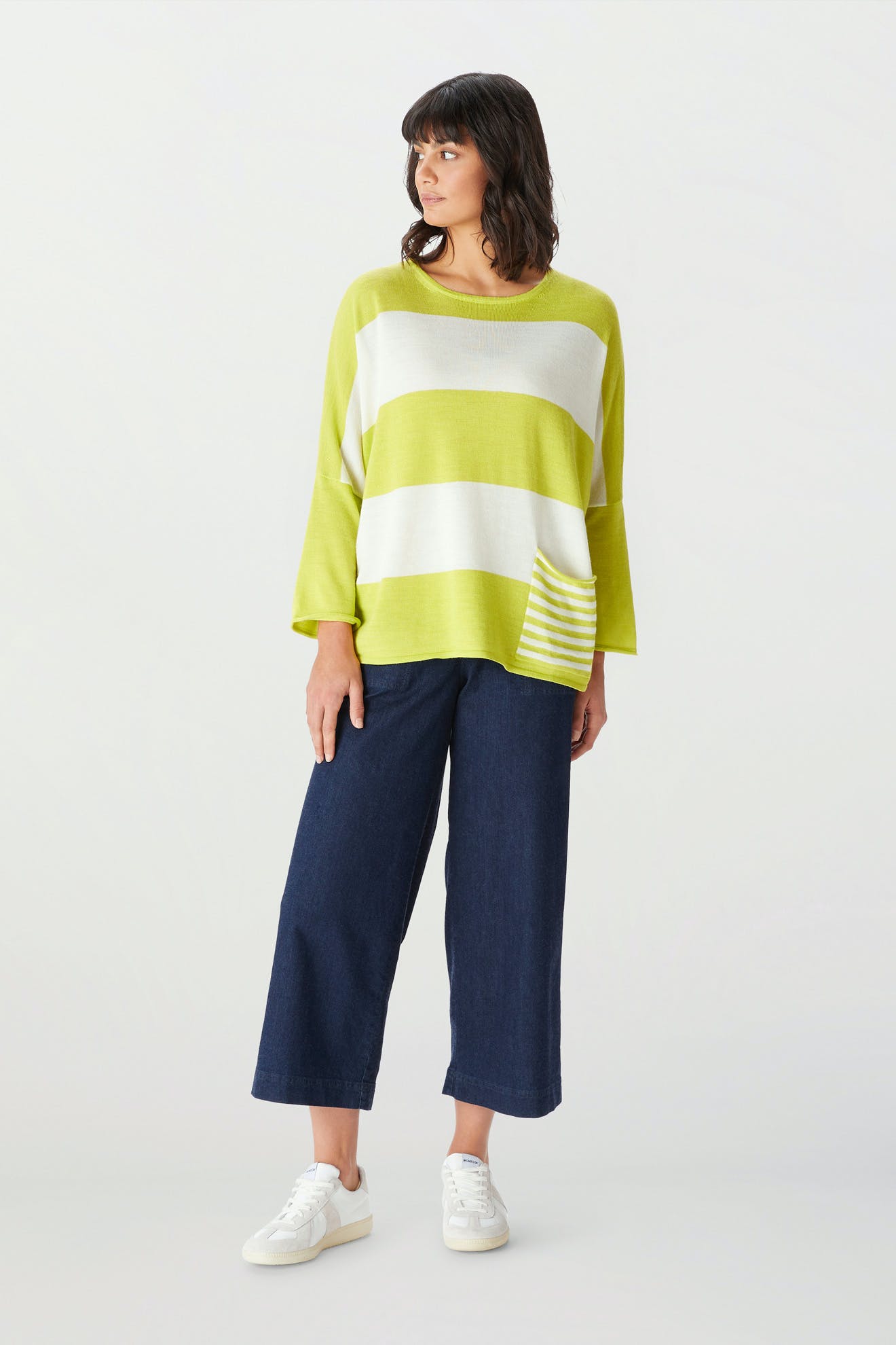 A woman wears the Merino Blend Stripe Sweater, featuring loose lime green and white stripes—ideal as a transitional weather piece—styled with wide-leg dark blue jeans and white sneakers, standing against a plain light background.