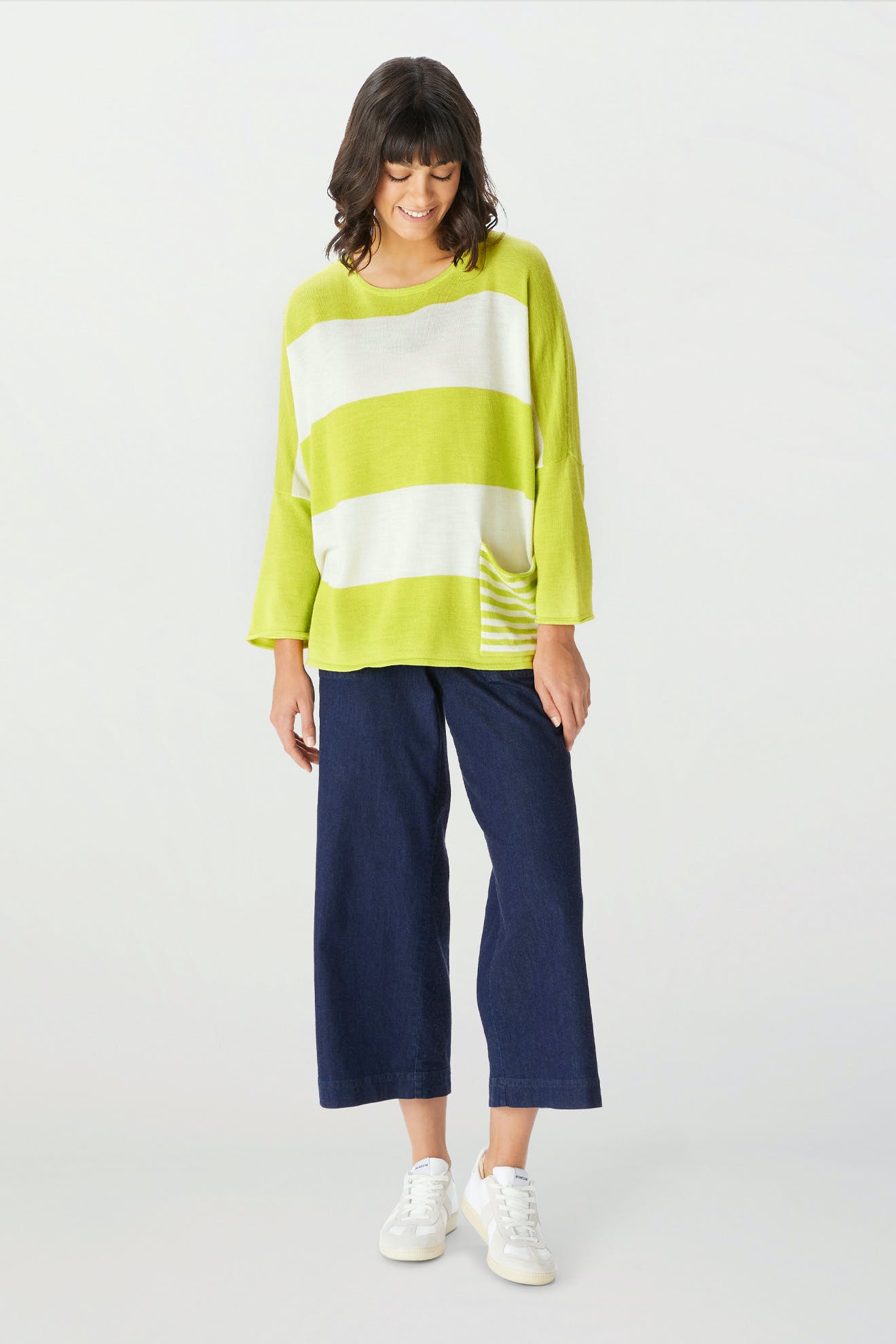 A woman stands against a plain light background, wearing the Merino Blend Stripe Sweater in loose lime green and white stripes—perfect for transitional weather—paired with wide-leg dark blue jeans and white sneakers.